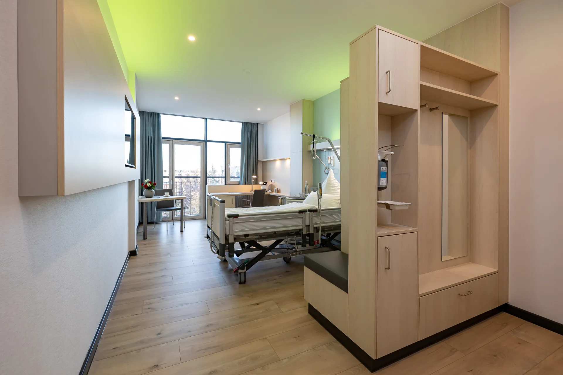 Modernly furnished patient room with a care bed, a bench seat, a cabinet and a large window with curtains