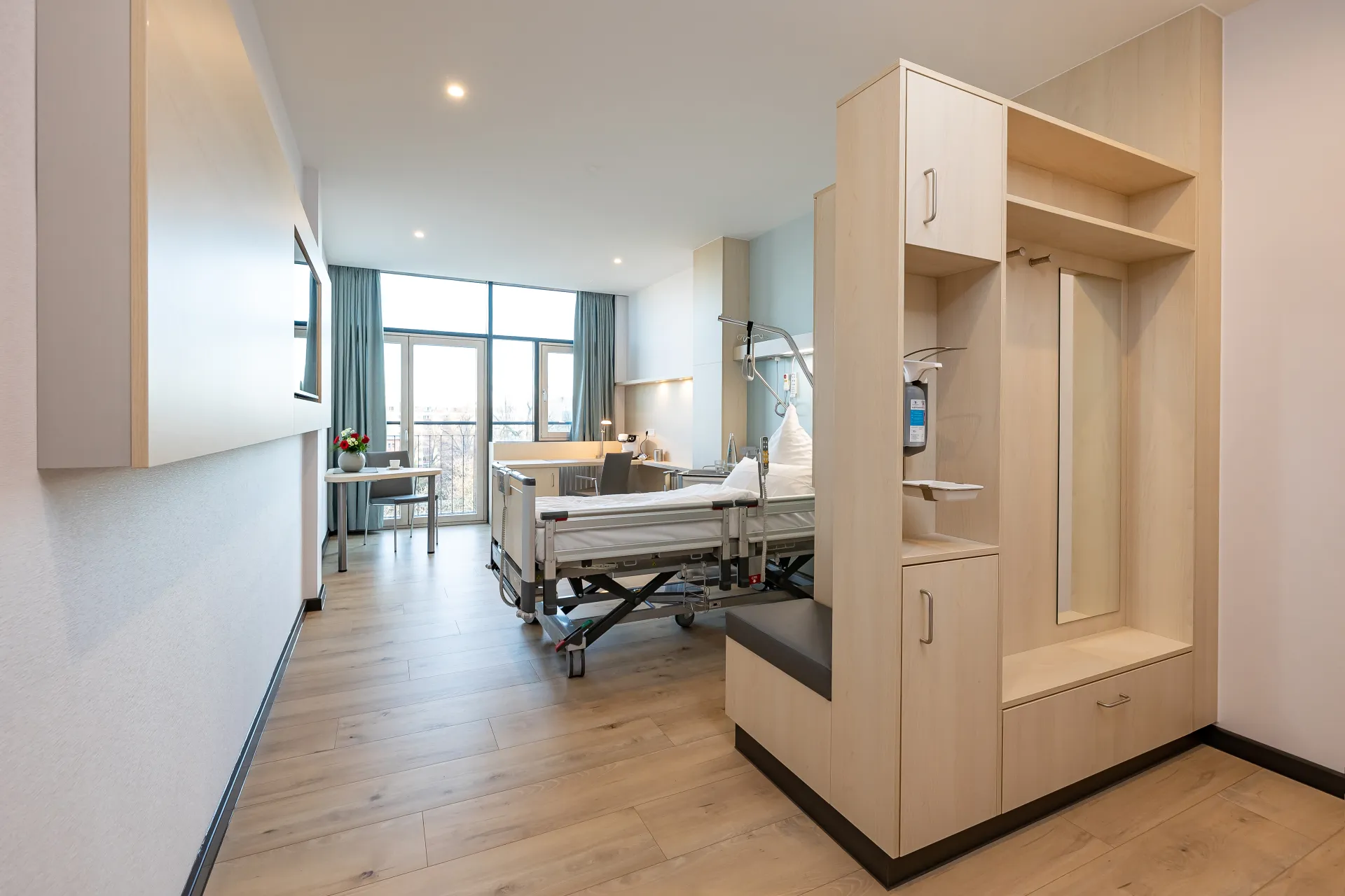 Modernly furnished patient room with a care bed, a window front, a desk and a wardrobe cabinet