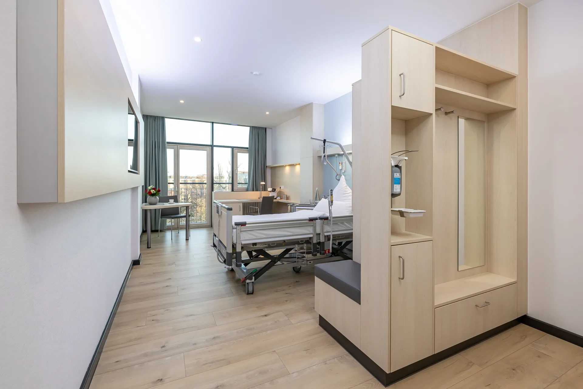 Bright patient room with a care bed, a bench seat, a coat rack and a large window with a view outside