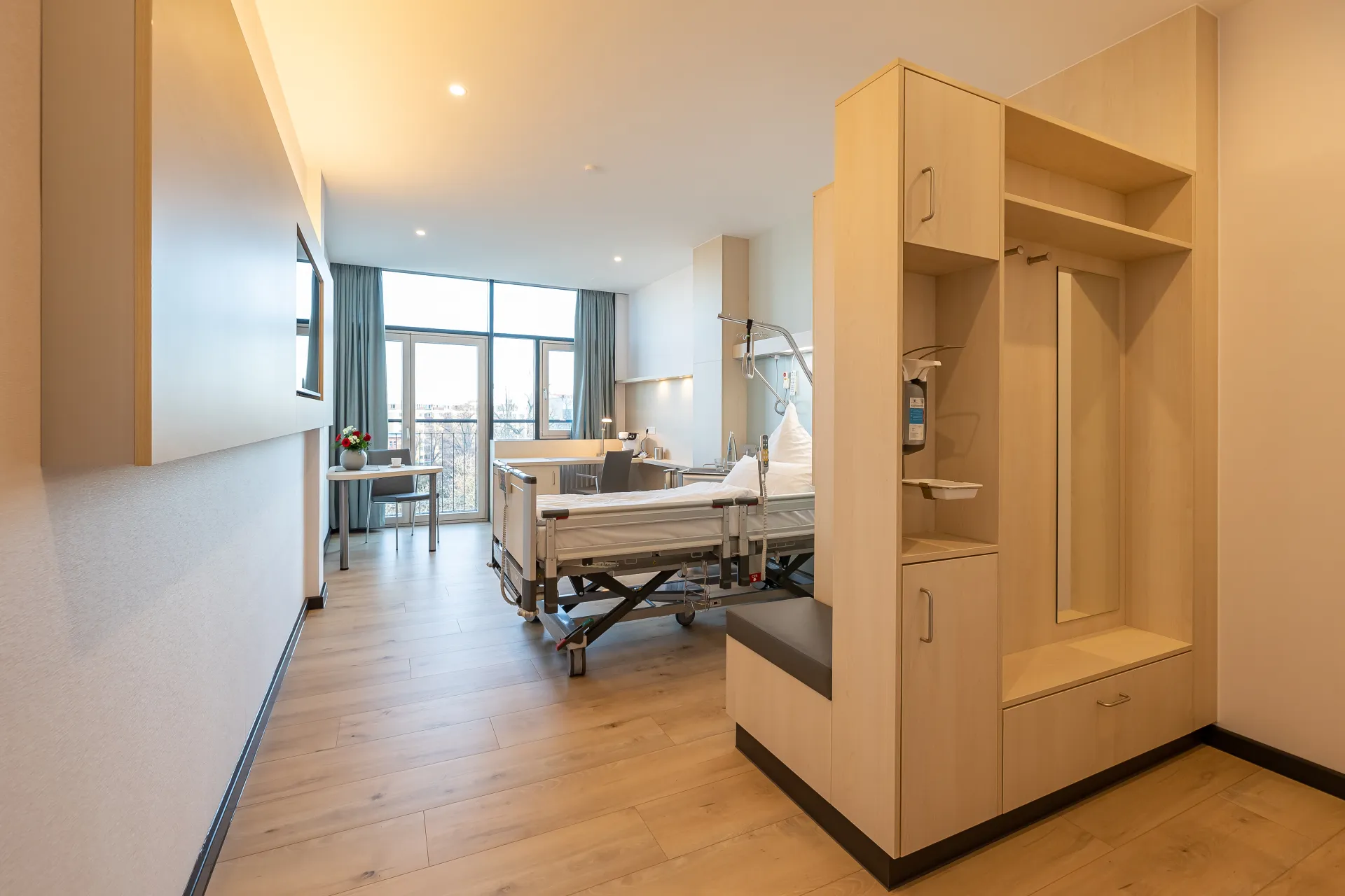 Bright patient room with a care bed, seating area, a large window with curtains, and an open wardrobe with a bench seat and shelves