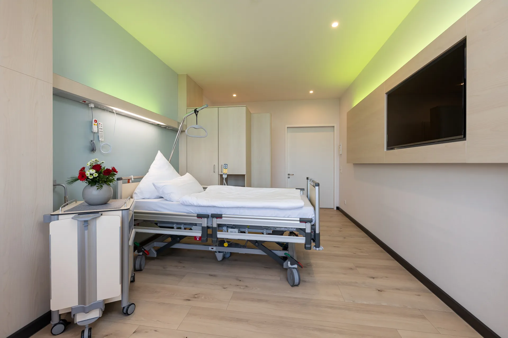Hospital room with empty bed, bedside table with a flower vase and a large wall-mounted television