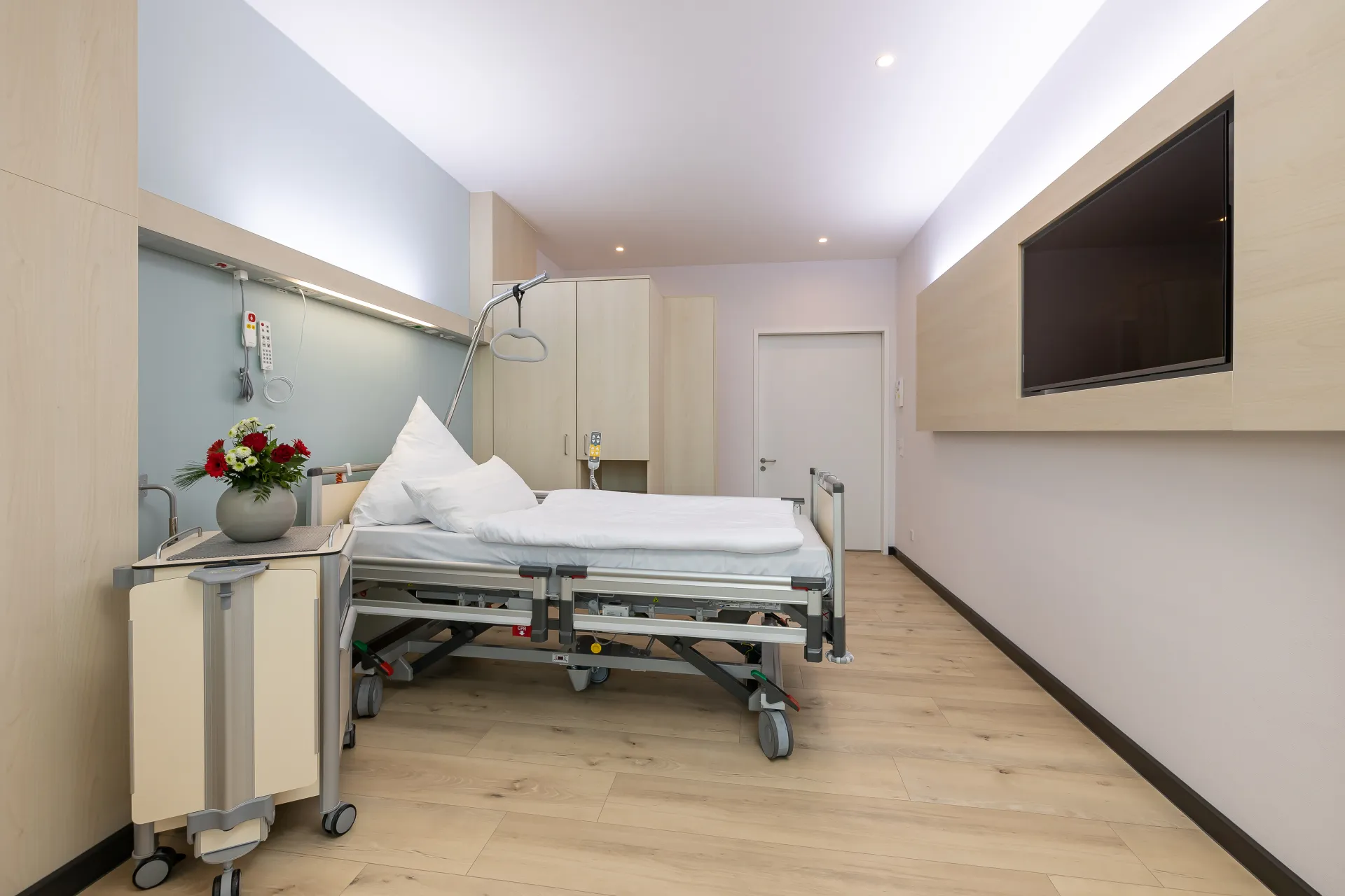 Hospital room with an empty bed, a bedside table with a vase and flowers, a large wall-mounted television and a wooden floor