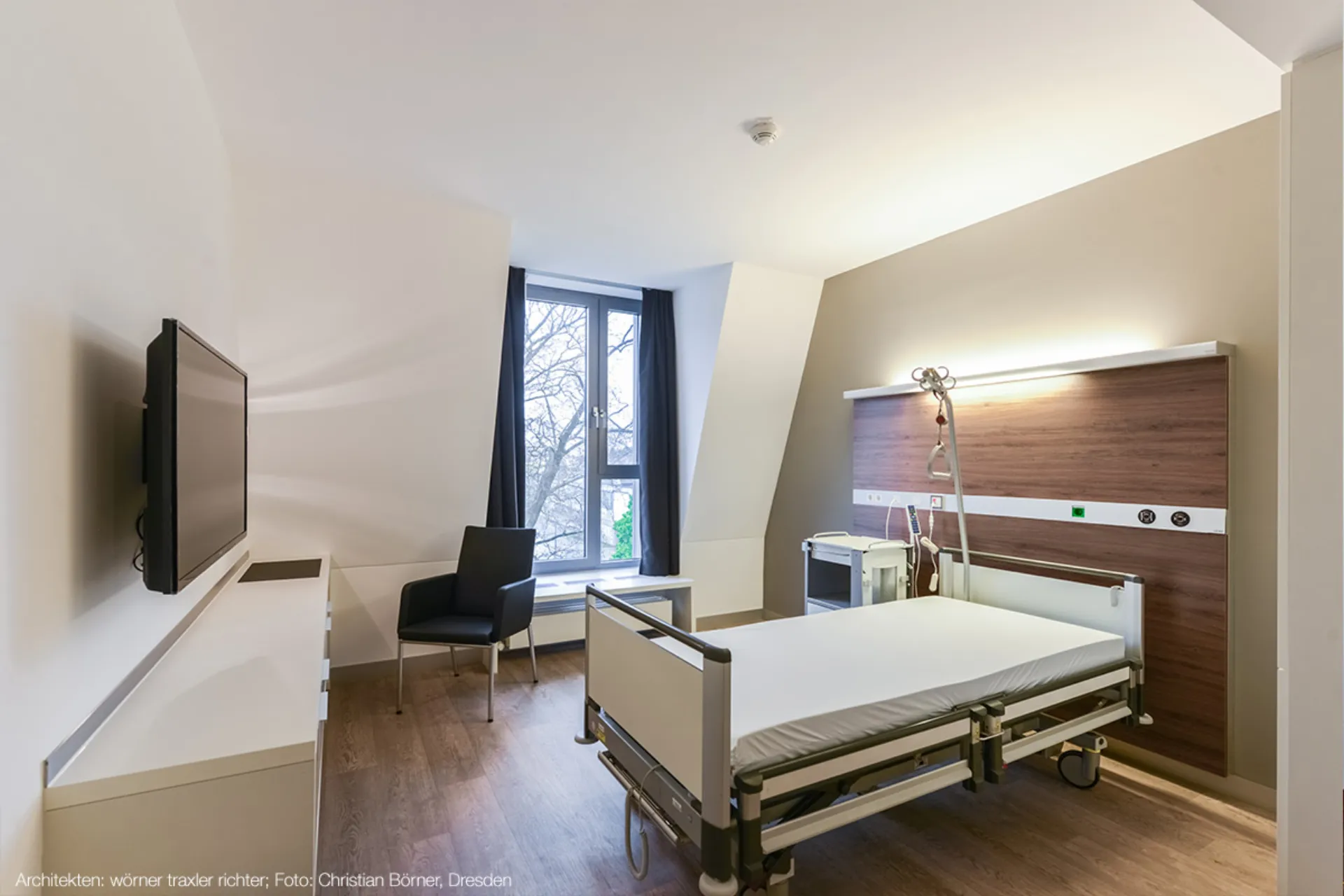 A simply furnished patient room with a hospital bed, a black chair, a window with curtains and a television on the wall