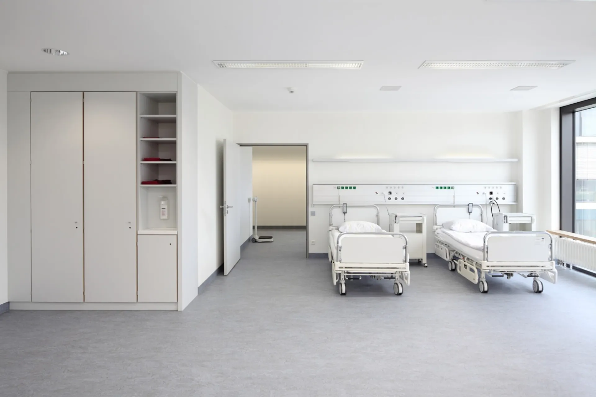 Two empty hospital beds with white bedding in a bright patient room with a cabinet and a large window
