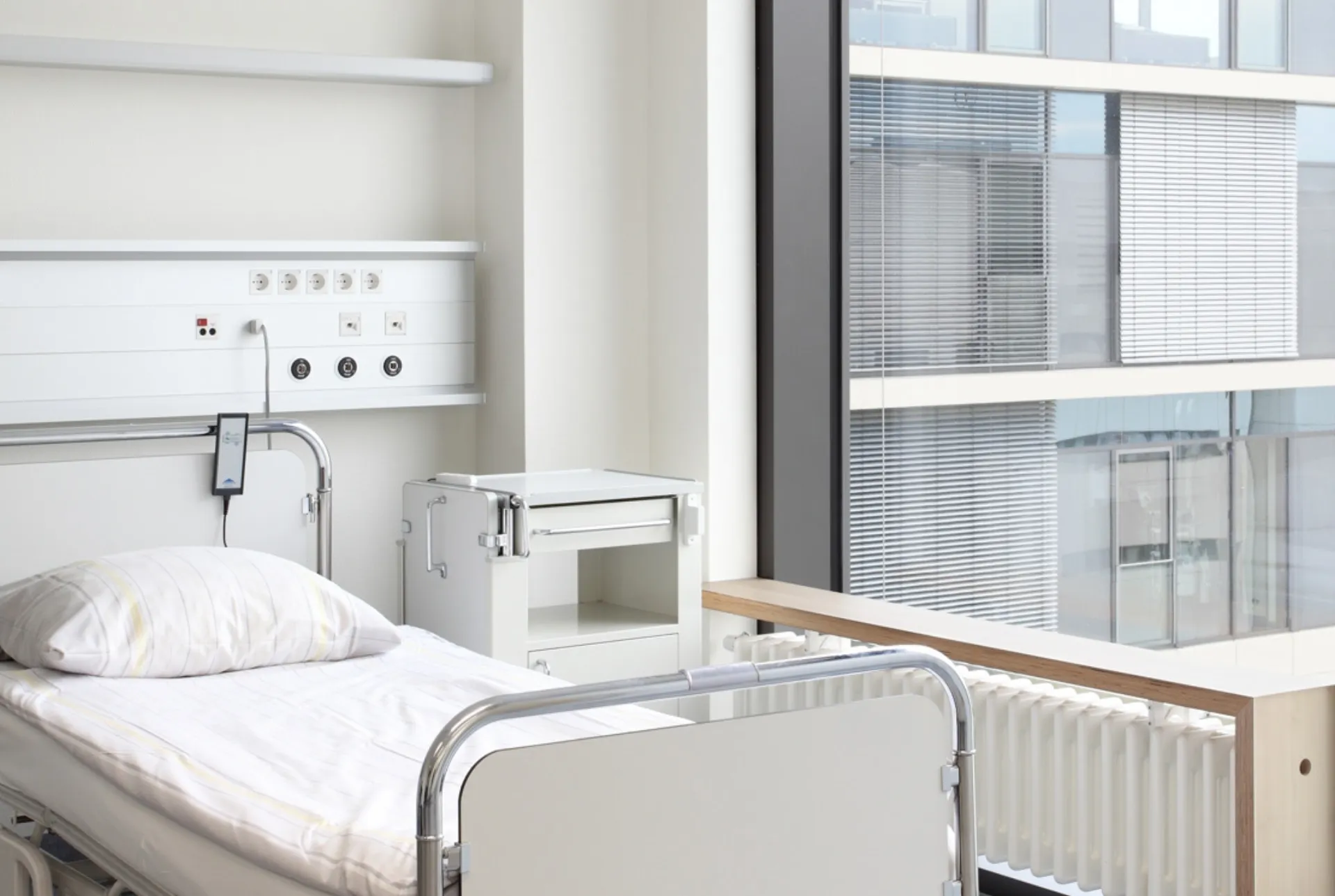 Hospital room with an empty bed, bedside table, medical wall console and a large window overlooking an office building