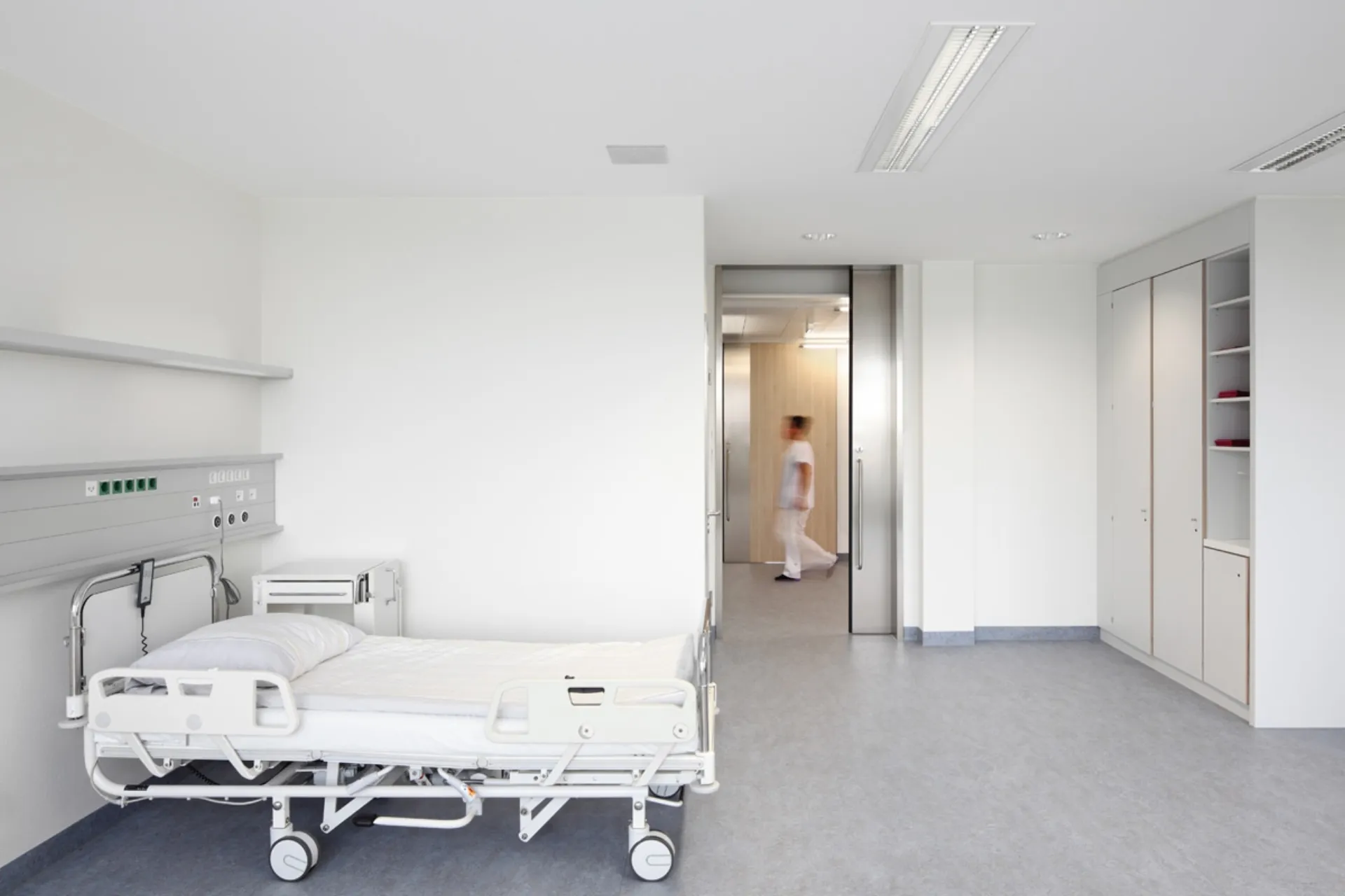 Empty hospital room with a bed on the left and a person in white clothing walking through a door in the background