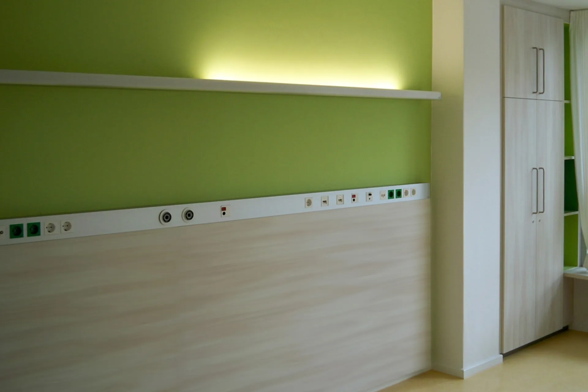 Wall with several sockets and connections beneath an illuminated white shelf in a room with a green-painted wall and a white cabinet