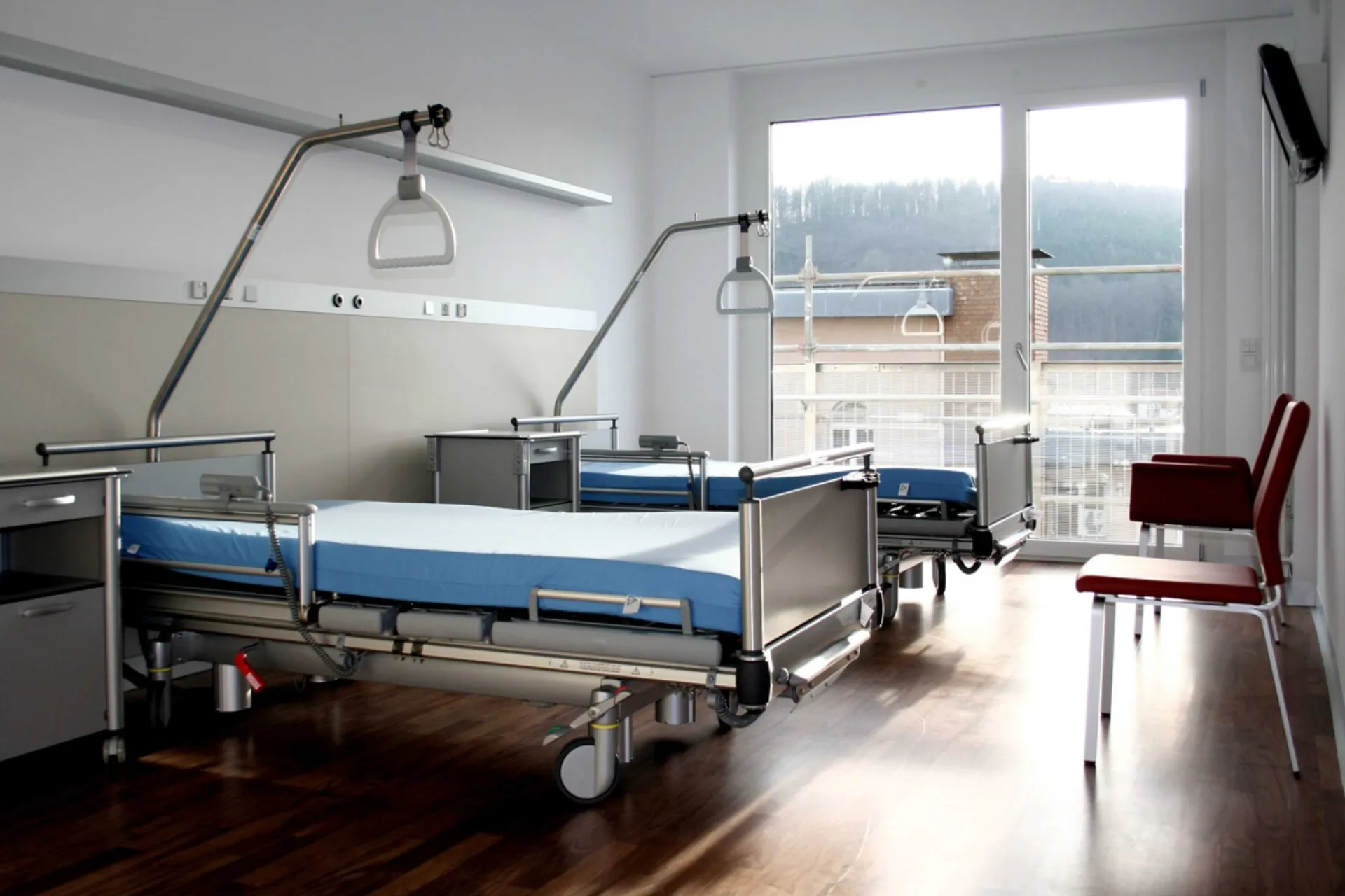 Empty hospital room with two beds, blue mattresses, two red chairs and a large window with a view of scaffolding and a wooded hill