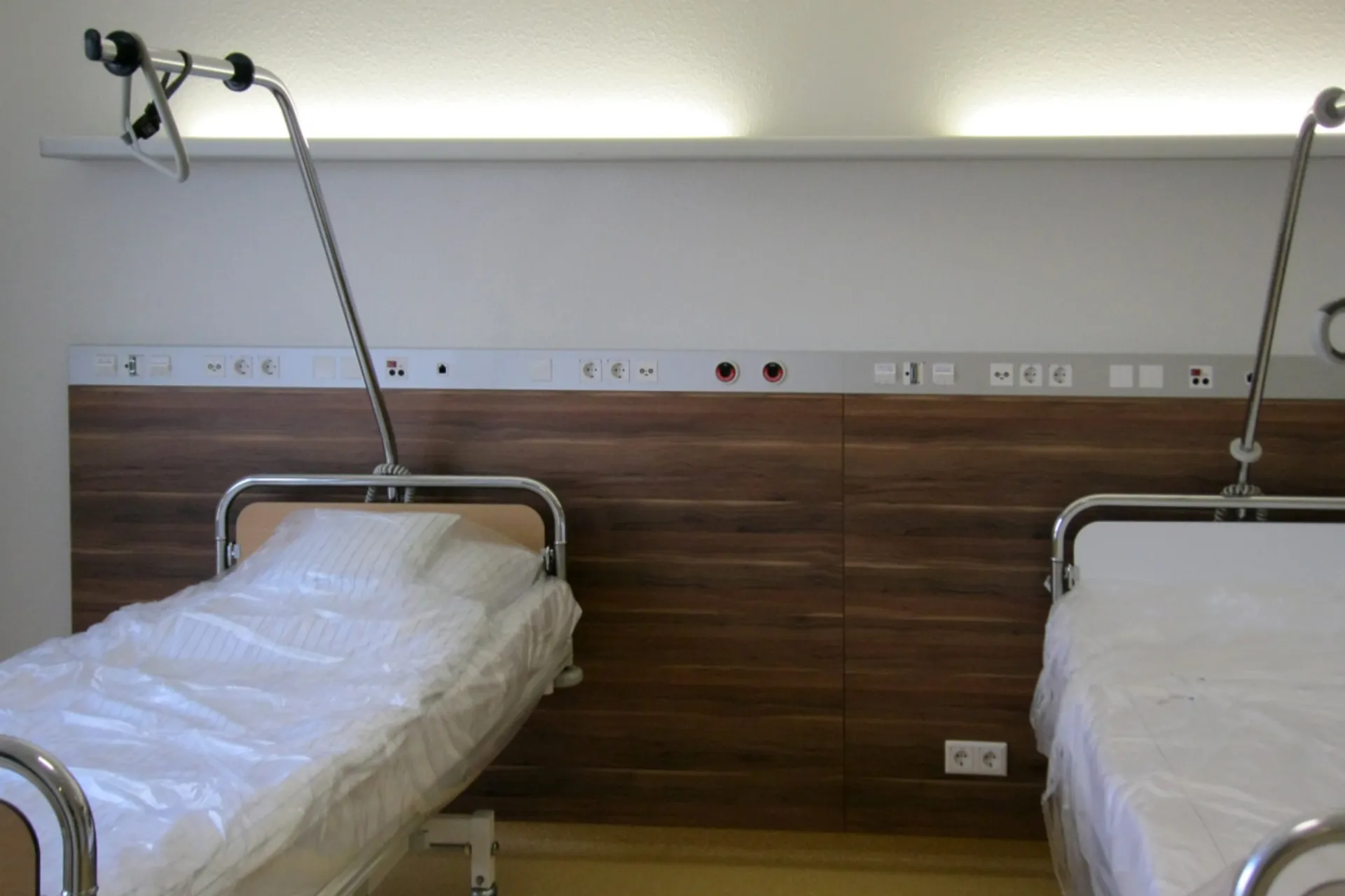 Two hospital beds with white covers and sitting-up aids in front of a wall with wooden cladding and medical connections
