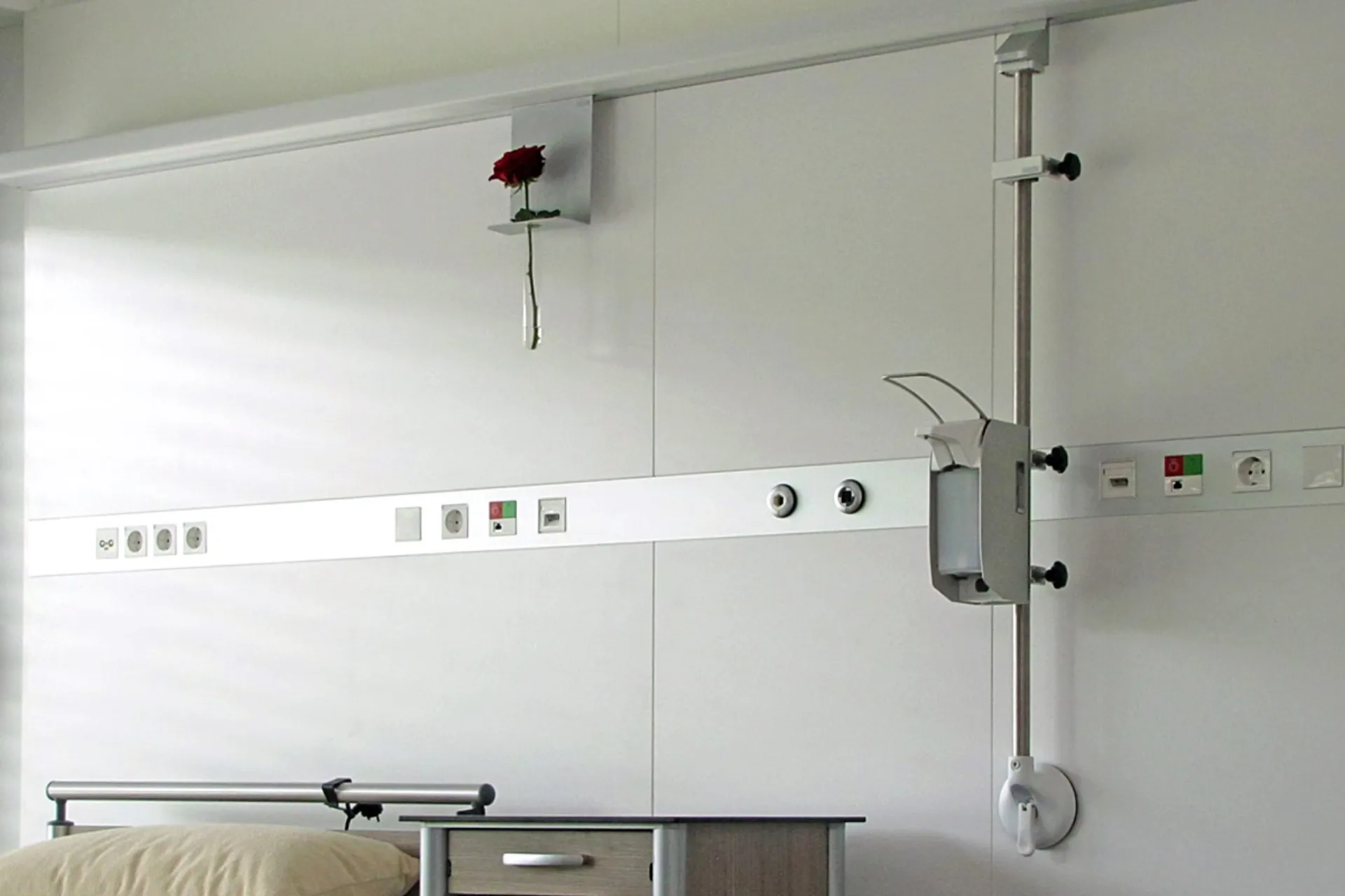 A flower vase holder and a rail with a hand sanitiser dispenser are hooked into the horizontal equipment rail on the rear wall of a patient room.