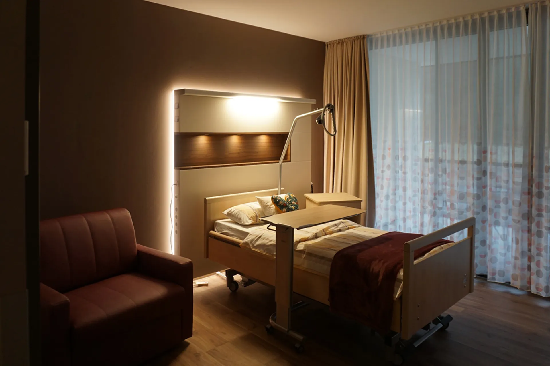 Krankenhauszimmer mit Pflegebett, Nachttisch, Leselampe, Sessel und großen Fenstern mit halbtransparenten Vorhängen
