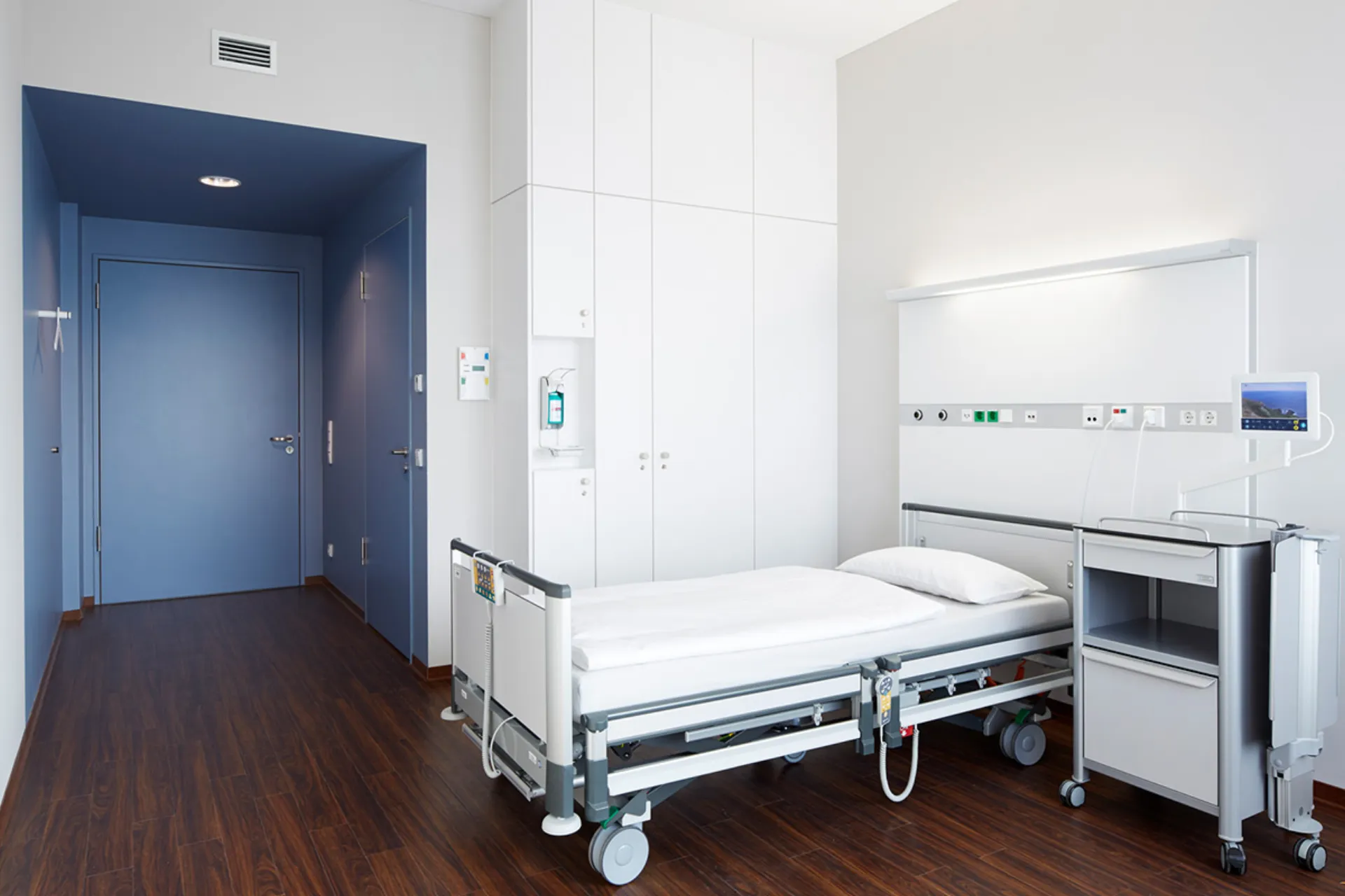 Hospital room with one bed, bedside table, white cabinets and a blue door in a bright space