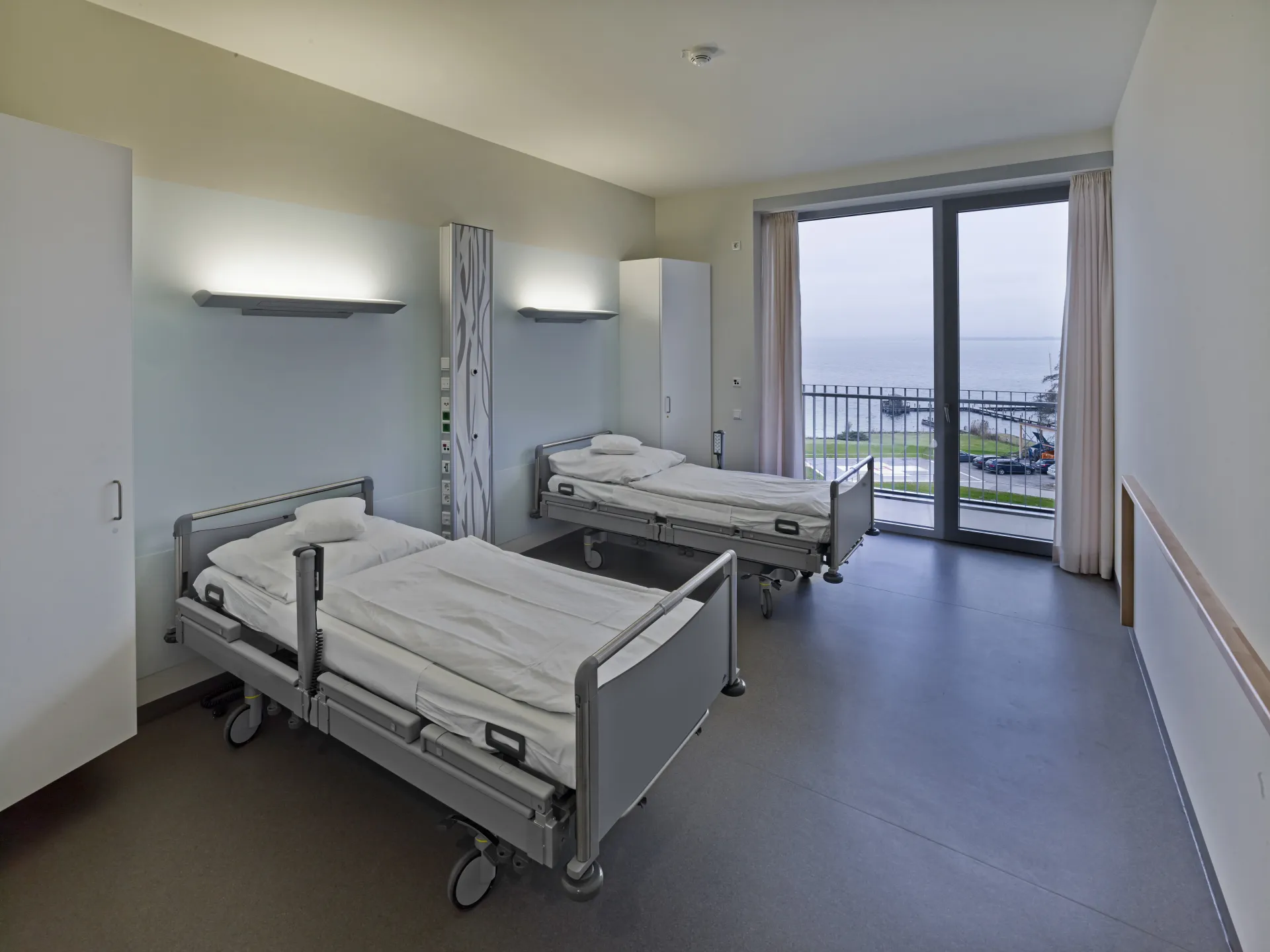 Krankenhauszimmer mit zwei Betten, Nachttisch, Schrank und großem Fenster mit Blick auf Wasser und Außenbereich