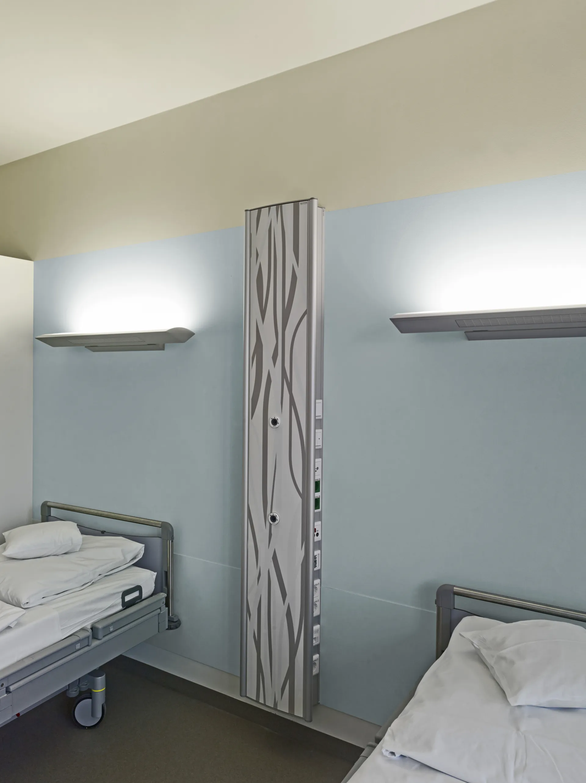 Zwei Krankenhausbetten mit weißen Bettdecken und Kissen an einer hellblauen Wand mit zwei Wandlampen und einer vertikalen Versorgungssäule mit grauem Muster