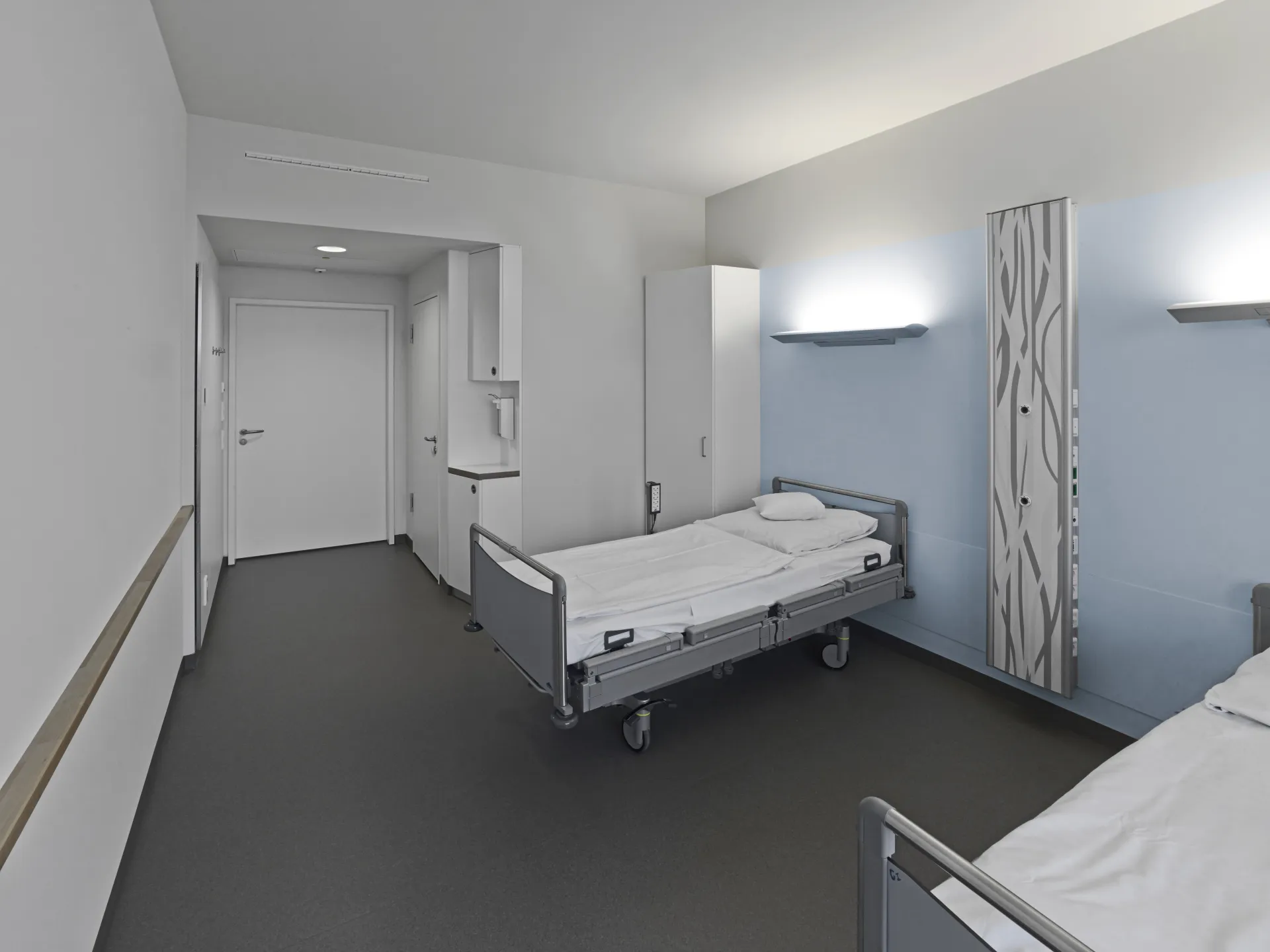 Zweibettzimmer mit Krankenhausbetten, weißen Wänden und blauer Wandverkleidung mit abstraktem Muster