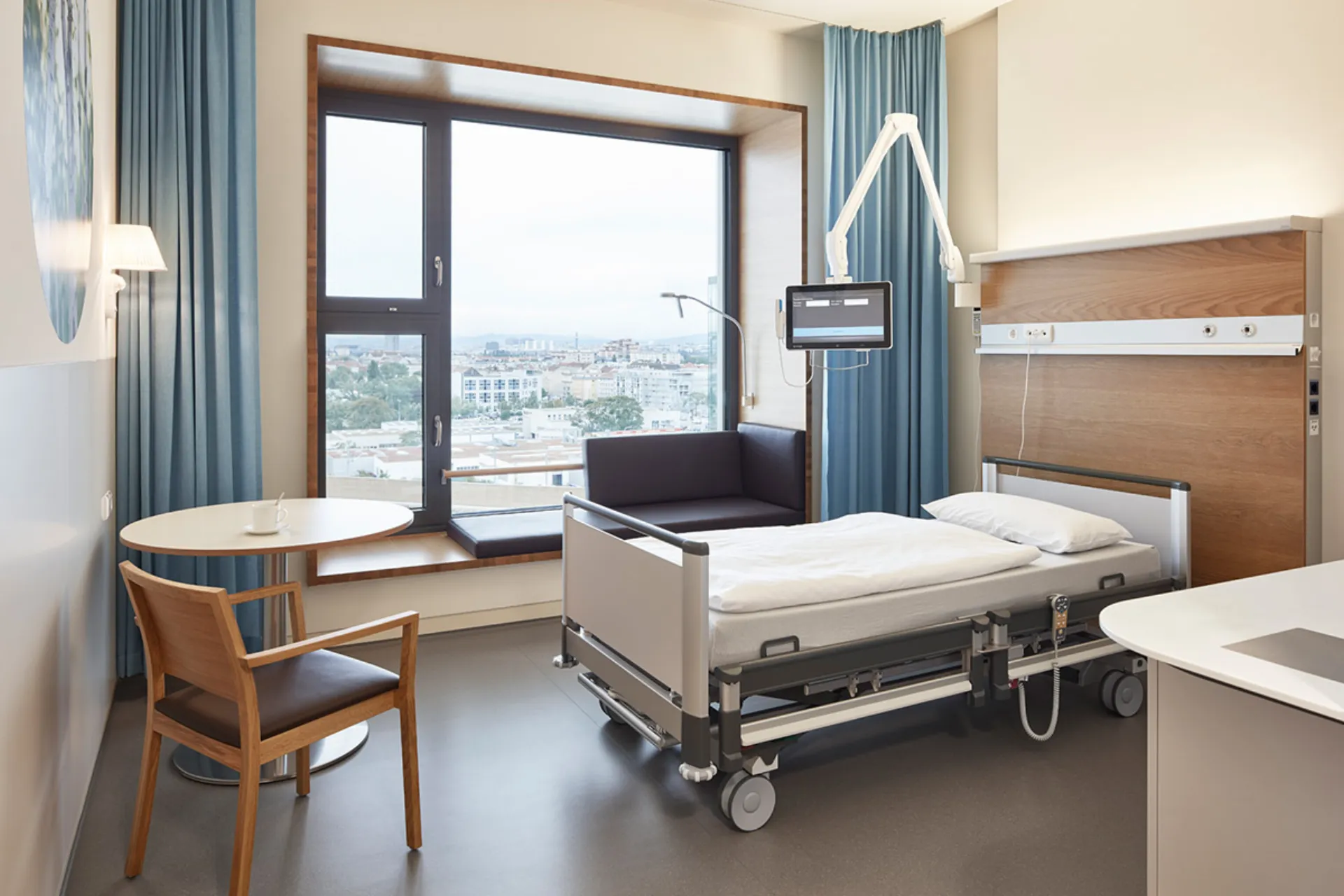 Hospital room with care bed, window seat with cushion, a chair and a small round table in front of a large window with a view of the city.