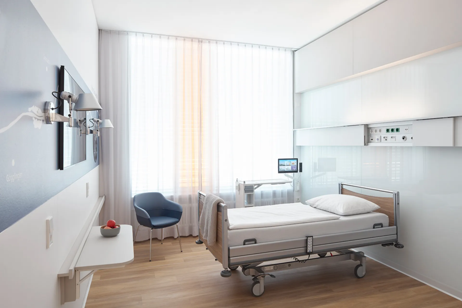 Modern furnished single room in a hospital with a care bed, blue chair and a large window with white curtains; the supply unit on the wall is slid open.