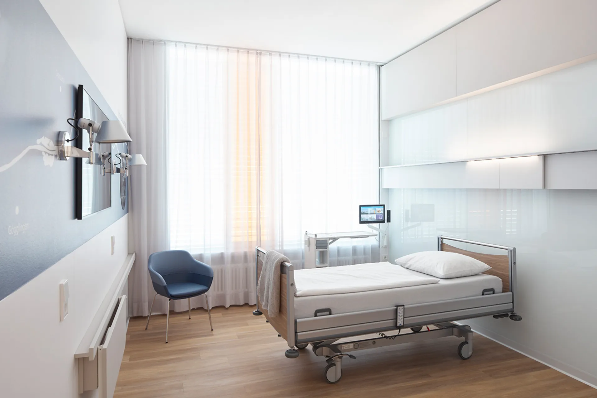 Modernly furnished single room in a hospital with a care bed, a blue chair and a large window with white curtains