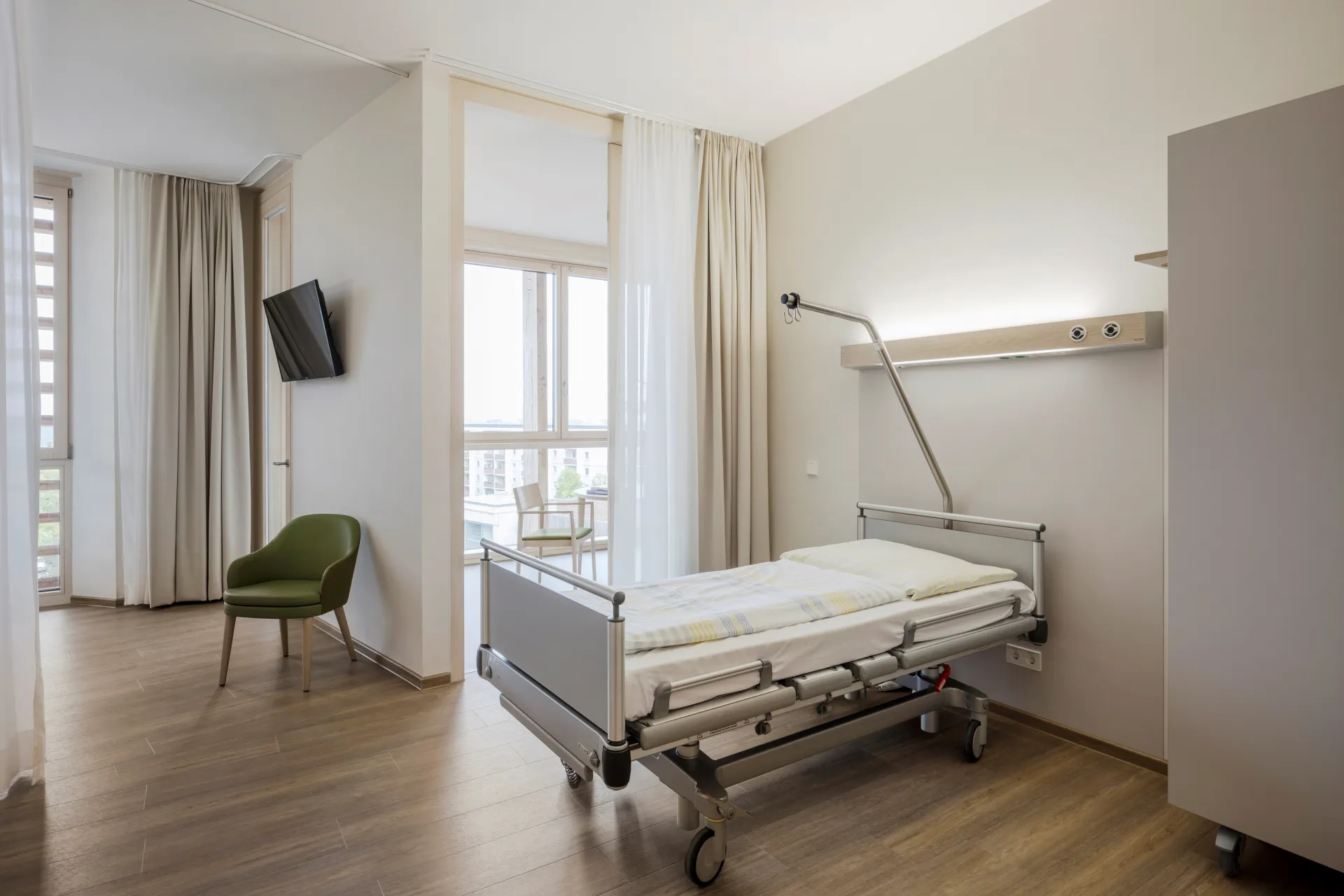 Hospital room with care bed, green chair, a window with a view of the city and a light wooden floor