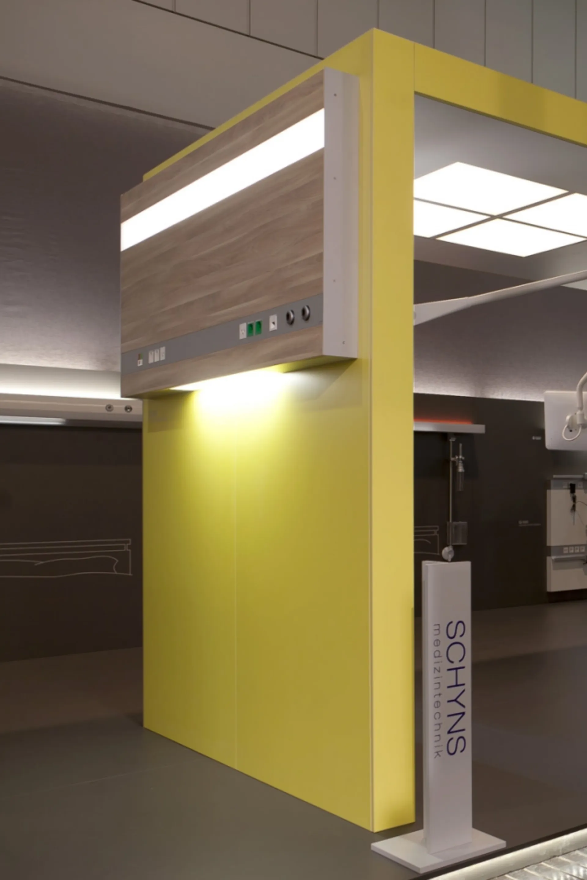 Yellow-painted wall with wood panel and integrated lighting in a showroom with a 'SCHYNS medizintechnik' display stand