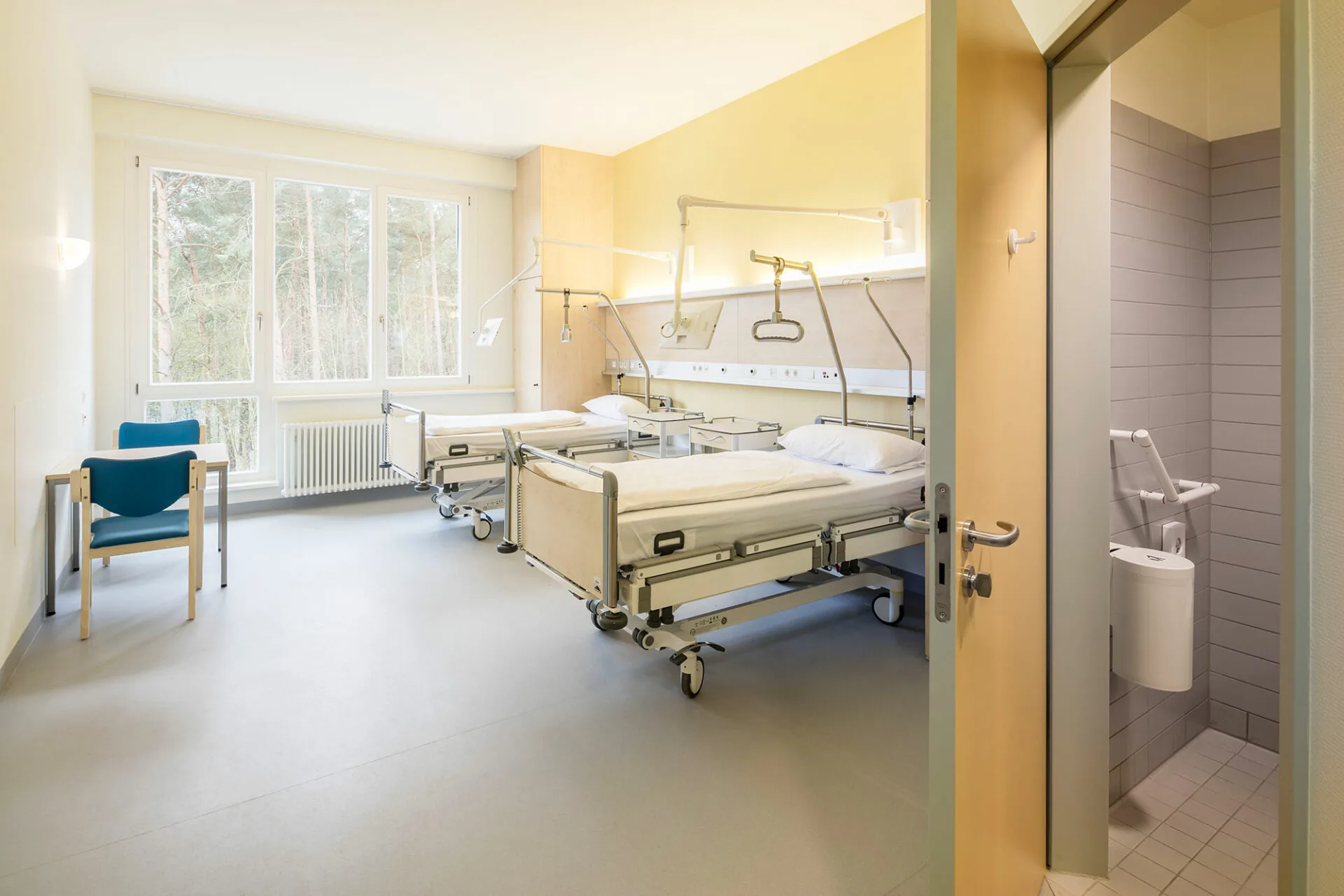 Twin room with a yellow accent wall, indirectly lit supply rail with swivelling reading lamps, a window with a forest view and an open door into the tiled bathroom