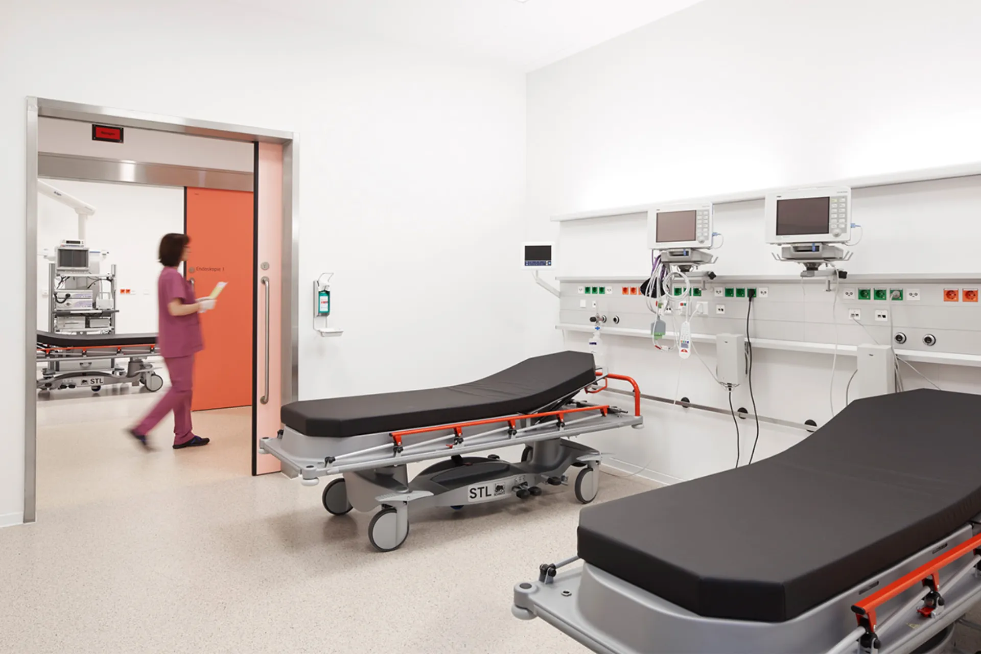 Treatment room with two couches, medical monitors on the wall and a person in medical clothing in front of an orange door
