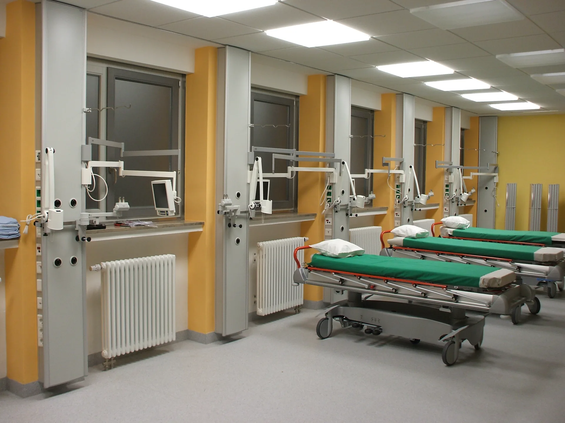 Aufwachraum mit mehreren Krankenliegen mit grünen Bettlaken, gelben Wandsäulen mit medizinischen Anschlüssen, Monitoren an Schwenkarmen und Untersuchungsleuchten