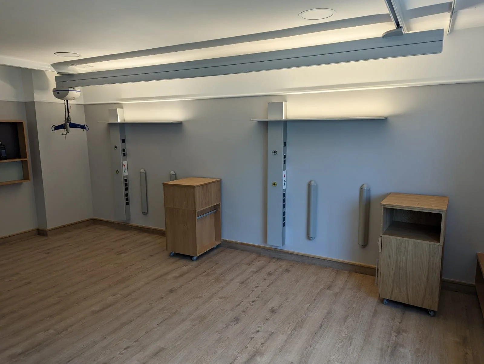 Two-bed area with two vertical pure supply columns on a grey wall, wooden bedside tables, indirect ceiling lighting and a patient lift