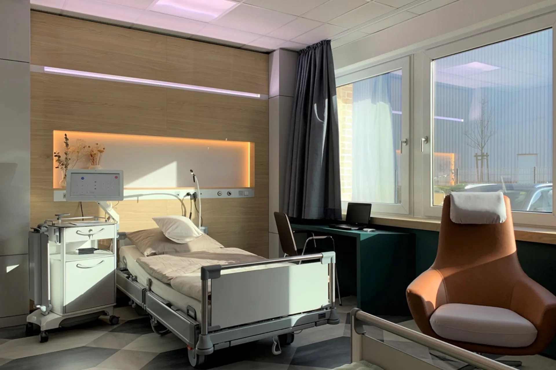 Krankenhauszimmer mit Pflegebett, medizinischem Gerät, Schreibtisch mit Stuhl am Fenster und einem gepolsterten Sessel