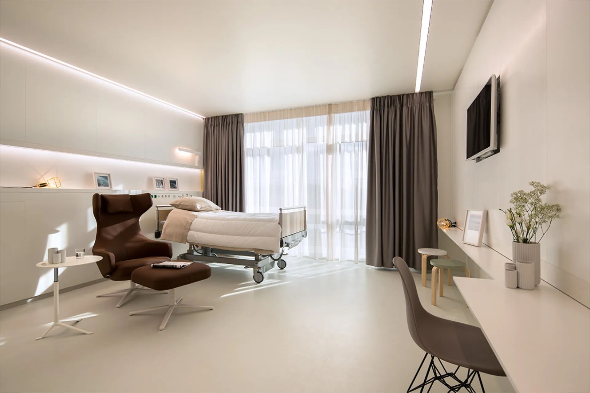 Bright, modern patient room with care bed, brown armchair with footstool, desk and a large window with curtains