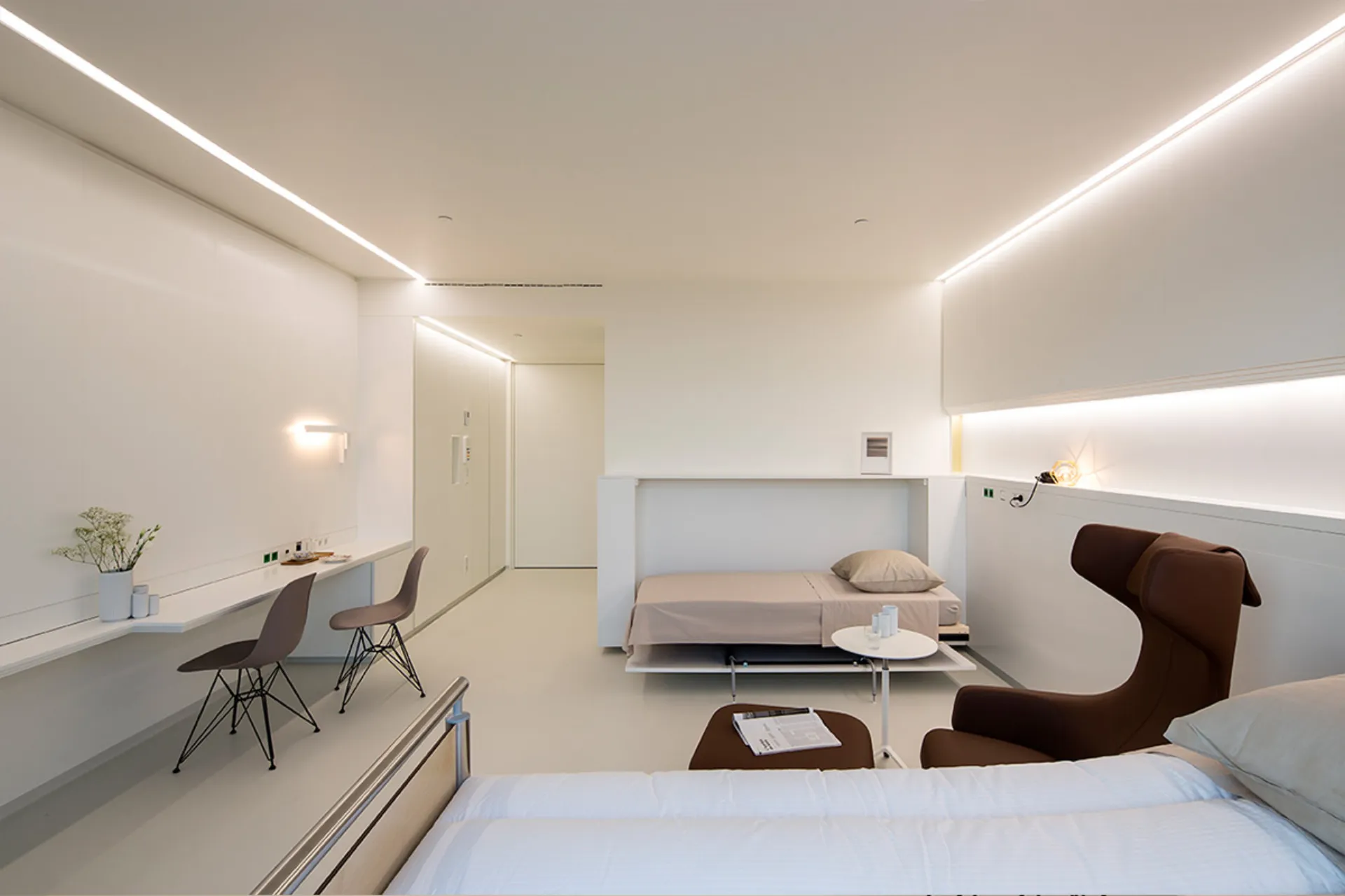 Minimally furnished white room with two beds, a brown armchair, a long table with two chairs and indirect lighting