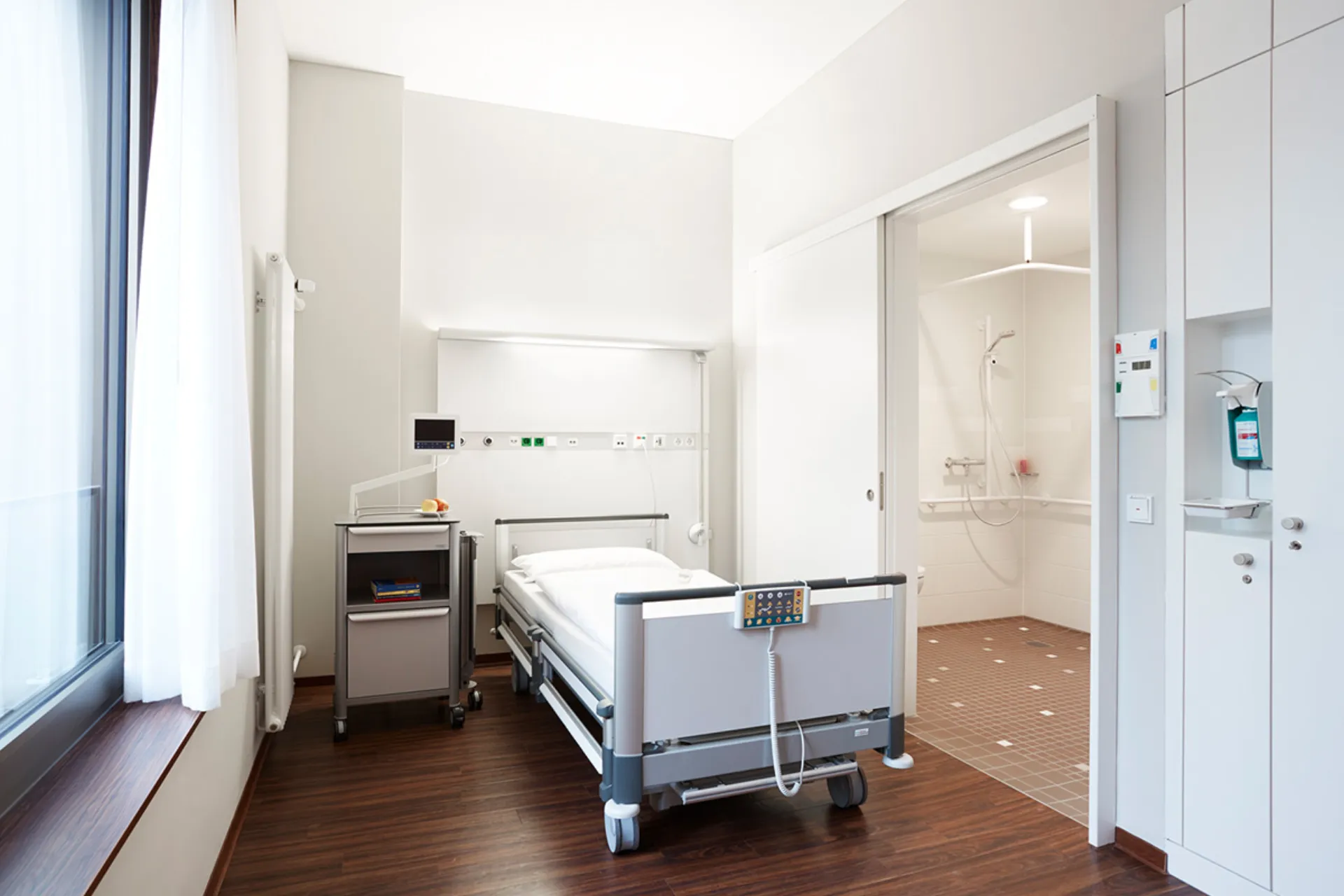 Hospital room with a bed, a grey duvet, a bedside table on castors and white cabinets on the wall