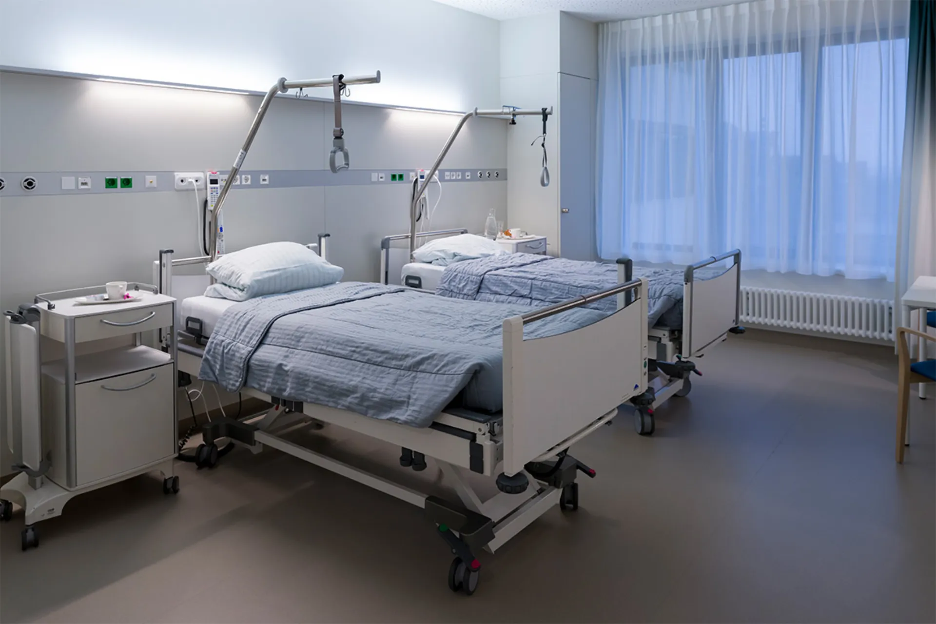 Two hospital beds with grey blankets in a bright patient room with a large window and curtains