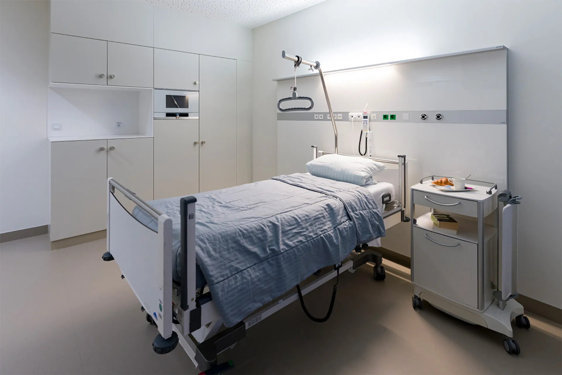 Hospital room with a bed, a grey duvet, a bedside table on castors and white cabinets on the wall