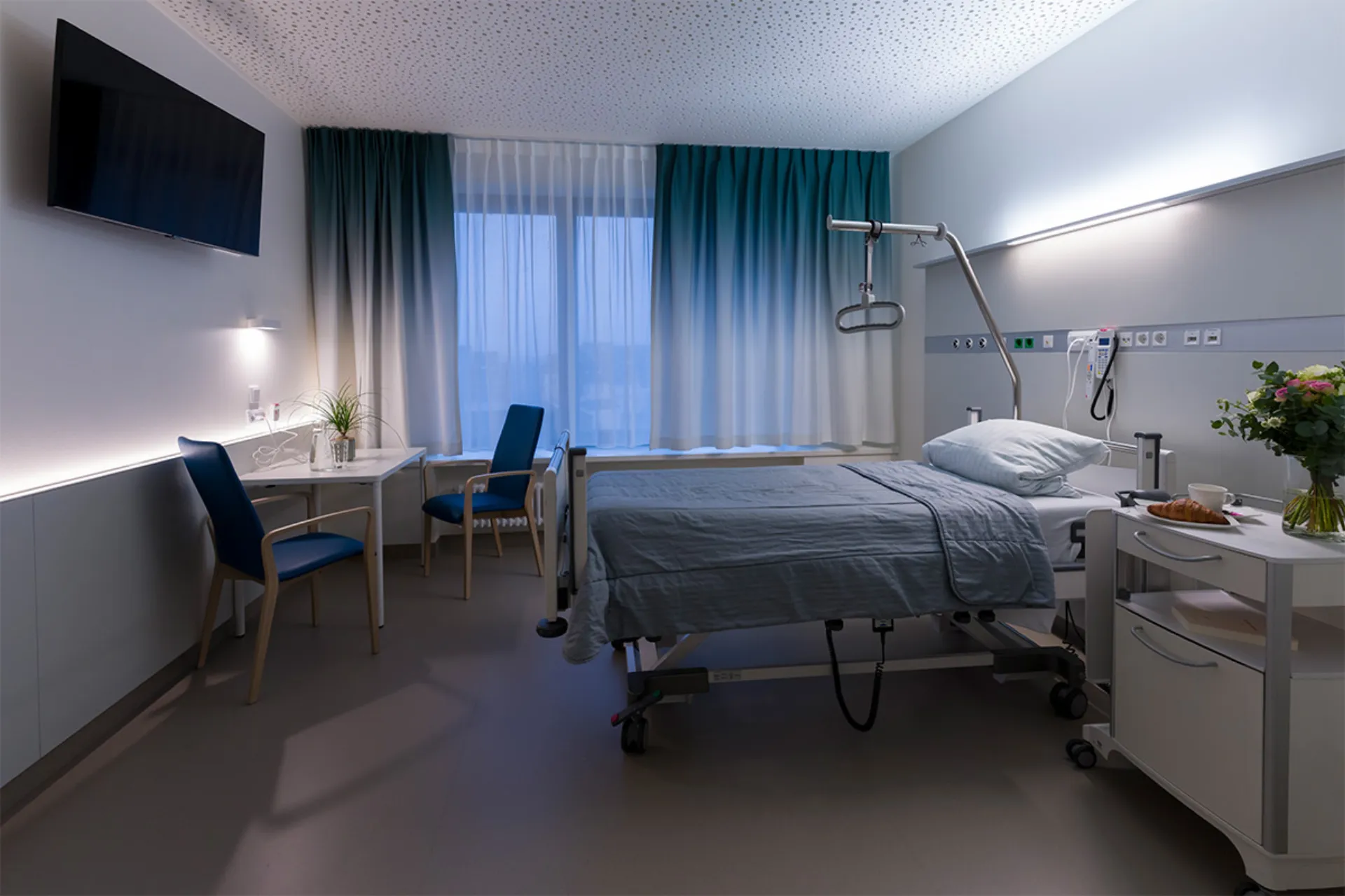 Hospital room with care bed, bedside table, two chairs, a desk, a large window with curtains and a wall-mounted television