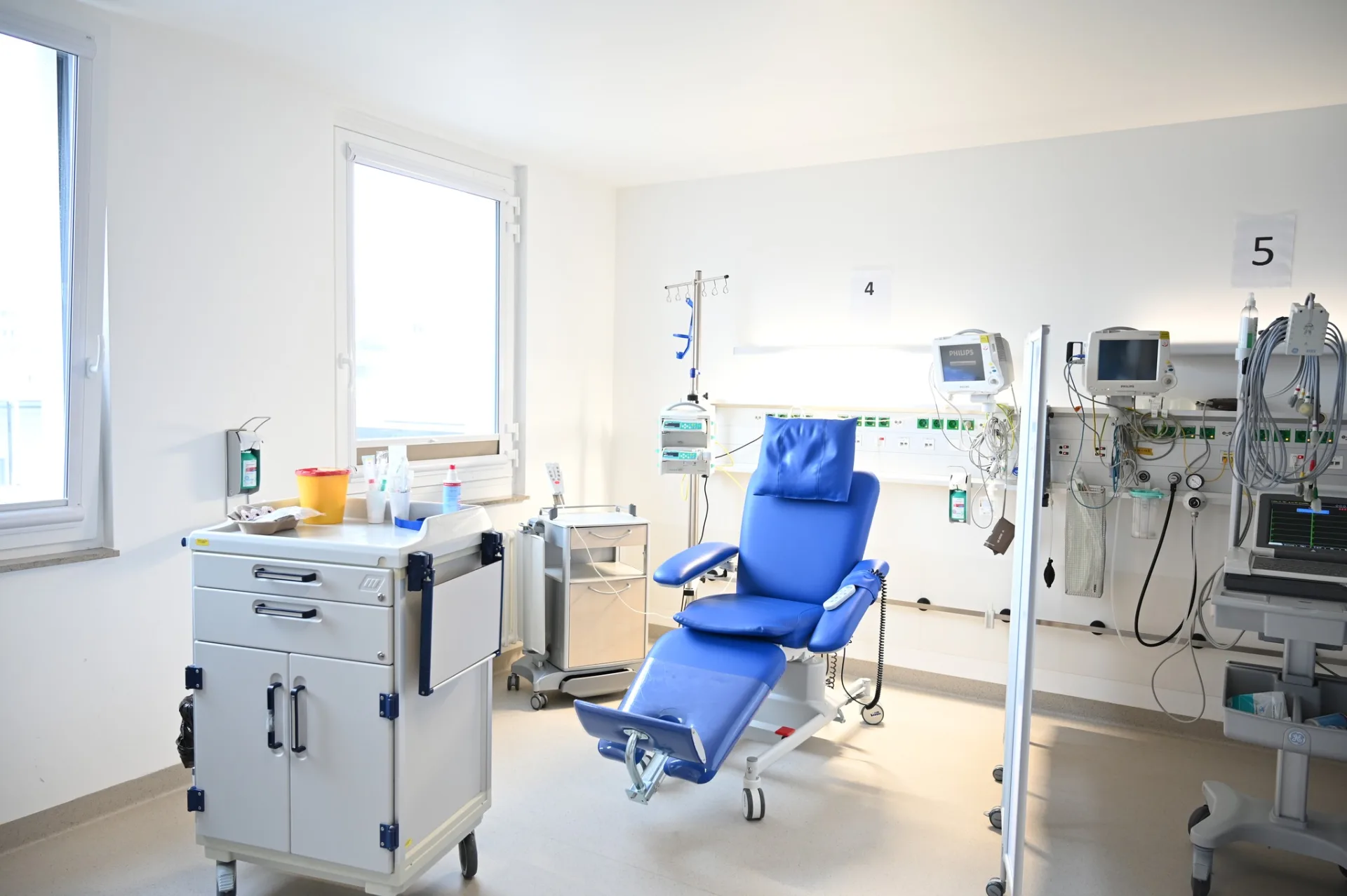 Dialysis station with a blue treatment chair, a white supply trolley, several patient monitoring screens on the wall and large windows with daylight