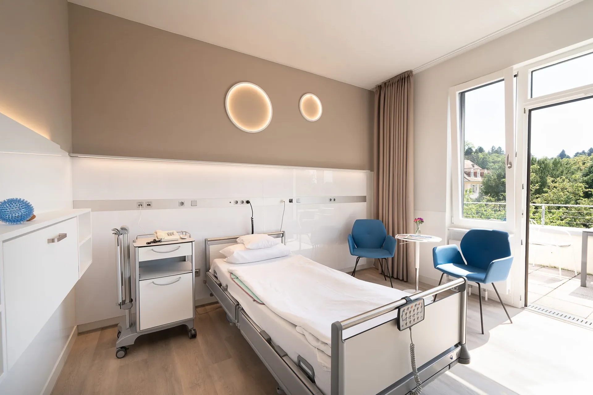 Modern patient room with care bed, bedside table on castors, two round wall luminaires, two blue armchairs by the side table and a balcony door with a view of greenery