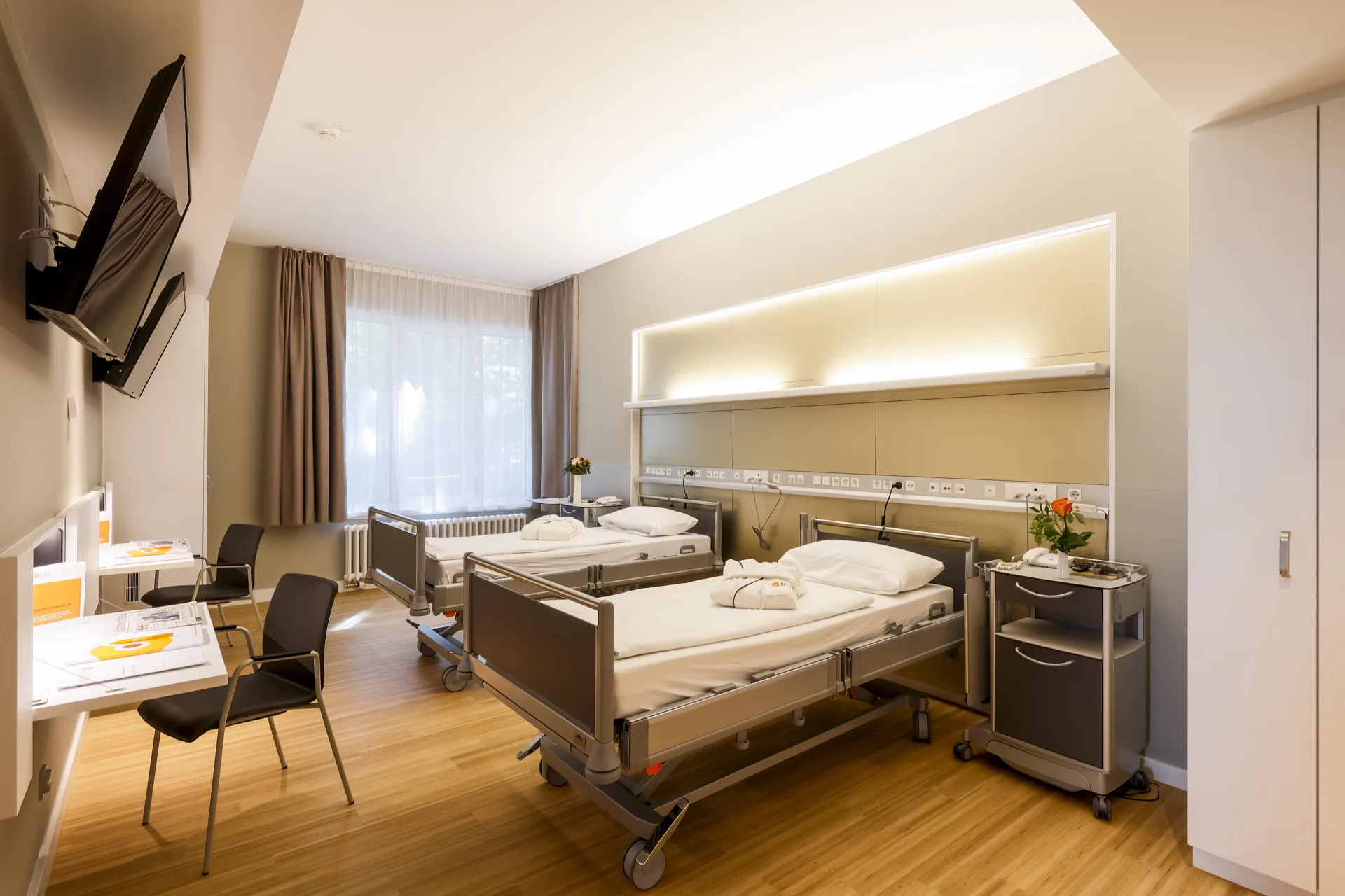 Patient room with two care beds, an indirectly lit supply strip on the rear wall, a desk with chair, a wall-mounted television and a window with a curtain