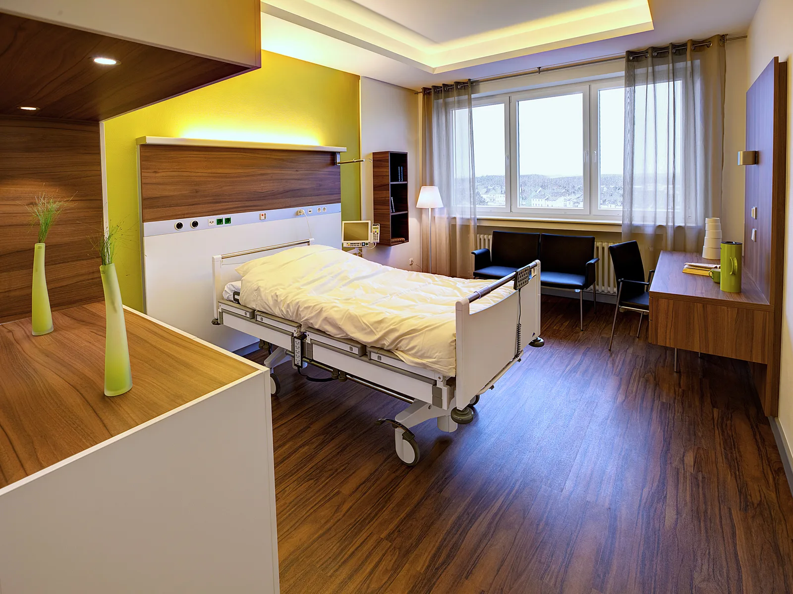 Patient room with care bed in front of a dark wood wall cladding, a supply rail backlit in yellow, a seating area with dark chairs and large windows with a view