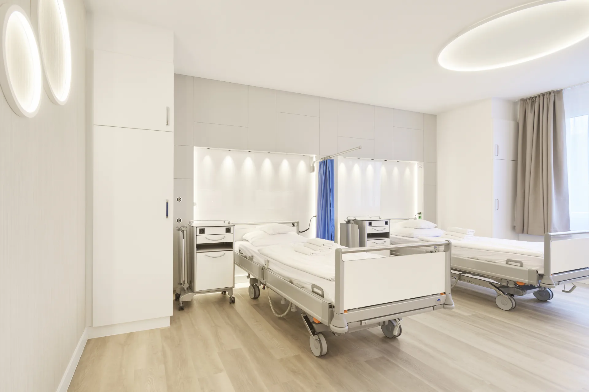 Zweibettzimmer mit Pflegebetten, runden Wand- und Deckenleuchten, indirekt beleuchteter Versorgungsnische und blauem Sichtschutzvorhang zwischen den Betten