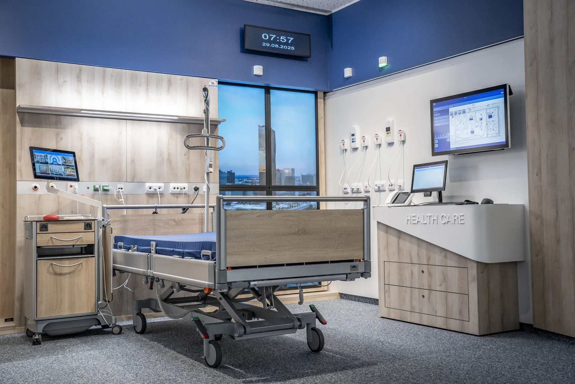 Modern intensive care room with care bed, blue accent wall, digital clock showing 07:57, two wall monitors, a care station with Healthcare lettering and a view of a skyline