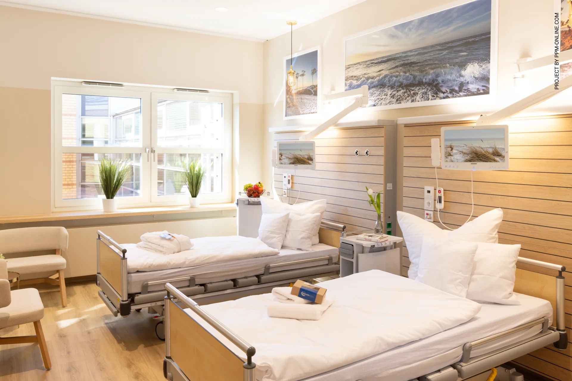 Helles Patientenzimmer mit zwei Pflegebetten, indirekt beleuchteter Versorgungsleiste, großformatigen Strandfotos an der Wand, Holzverkleidung und Topfpflanzen auf der Fensterbank