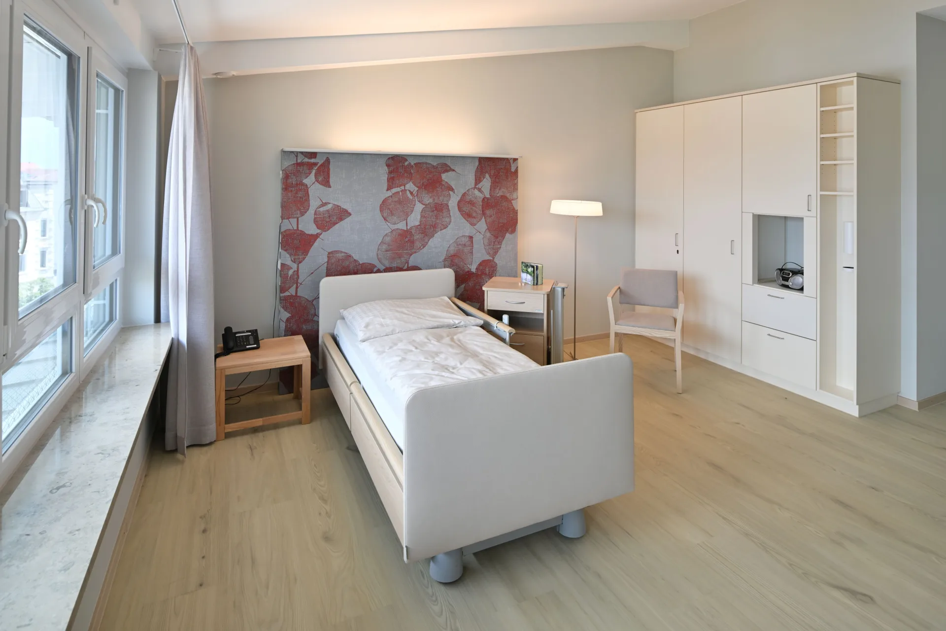 Großzügiges Patientenzimmer mit Pflegebett, rot-grauem Blattmotiv-Wandbild, weißem Schrank mit Fernsehnische, hellem Parkettboden und großen Fenstern