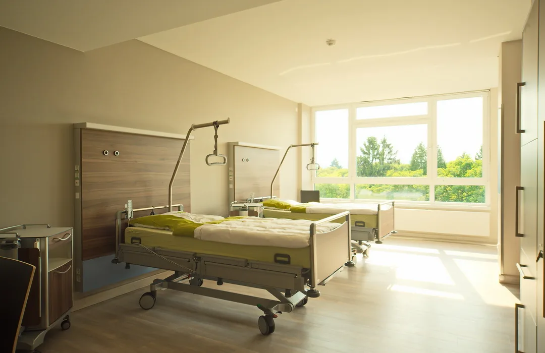 Patientenzimmer mit zwei Pflegebetten, Patientenlifter-Tragebügeln über den Betten, großem Fenster mit Blick ins Grüne und hellem Holzboden