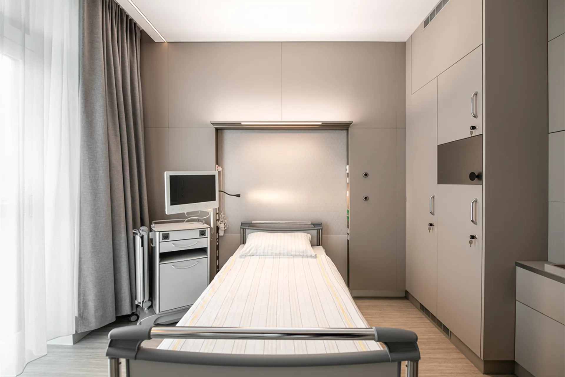 Krankenhauszimmer mit Pflegebett, Nachttisch, zwei Stühlen, Schreibtisch, großem Fenster mit Vorhängen und Wandfernseher