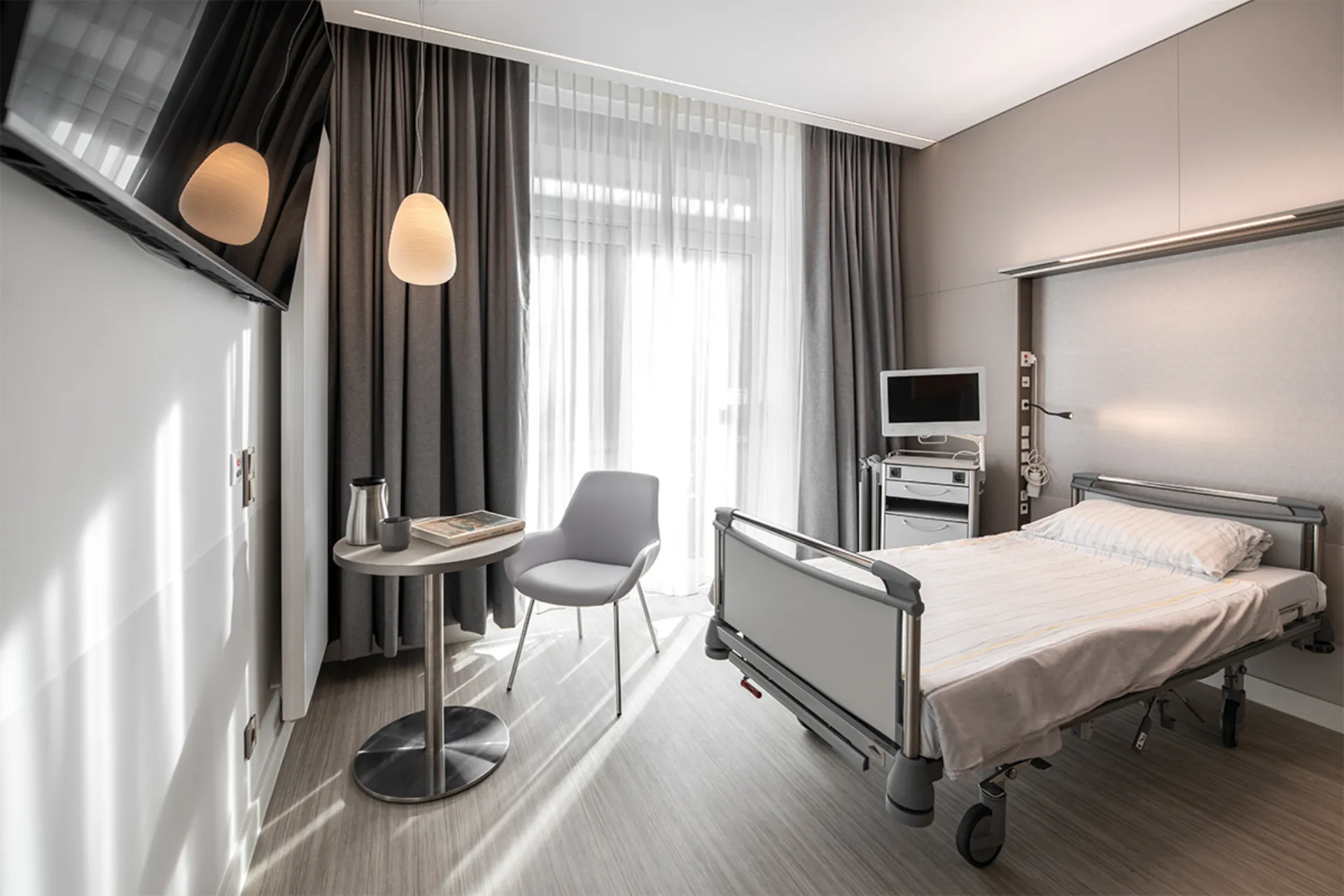 Krankenhauszimmer mit Einzelbett, Nachttisch, Stuhl, Fernseher und großen Fenstern mit Vorhängen