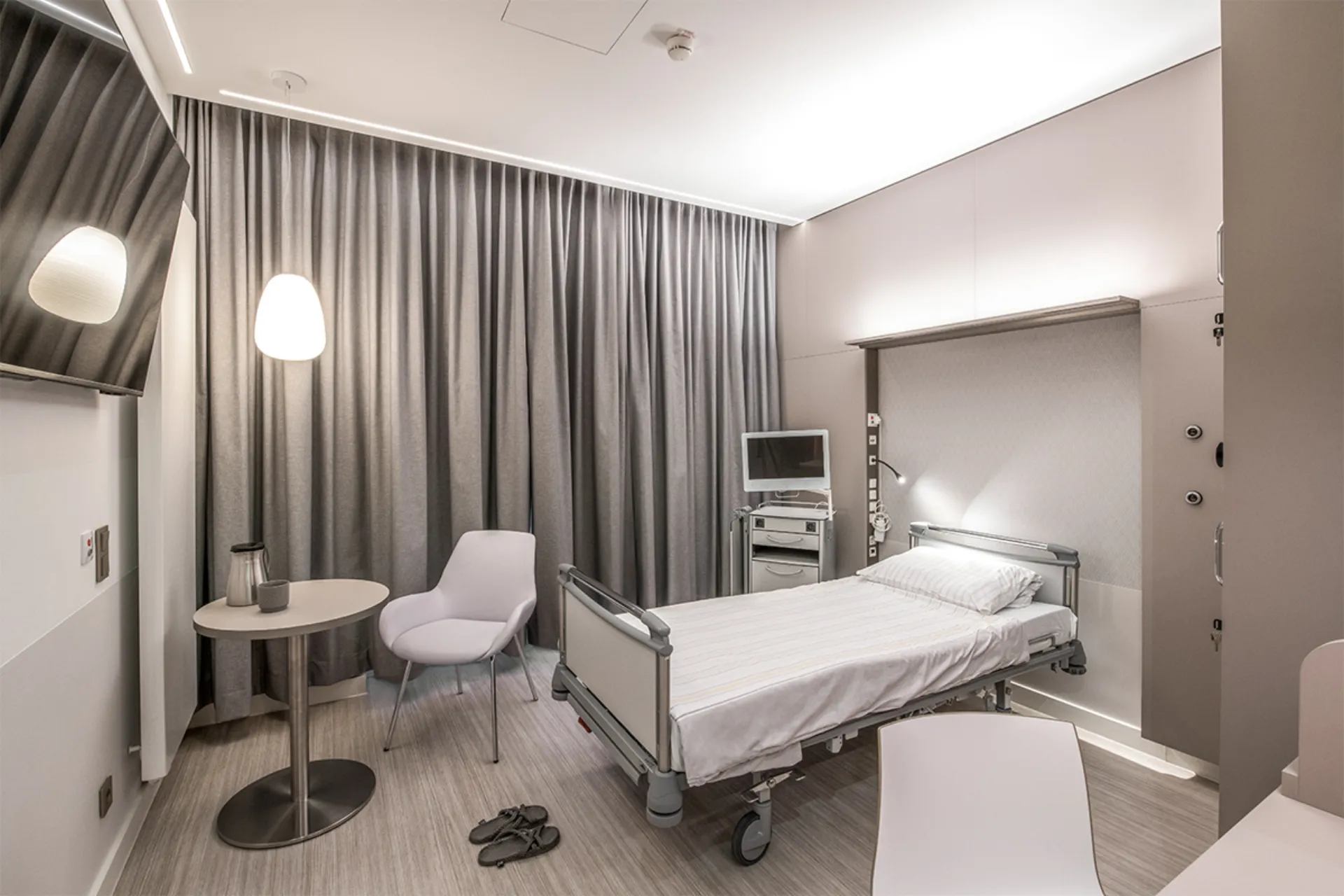 Krankenhauszimmer mit einem Bett, einem runden Tisch, zwei Stühlen, Vorhängen und Hausschuhen auf dem Boden