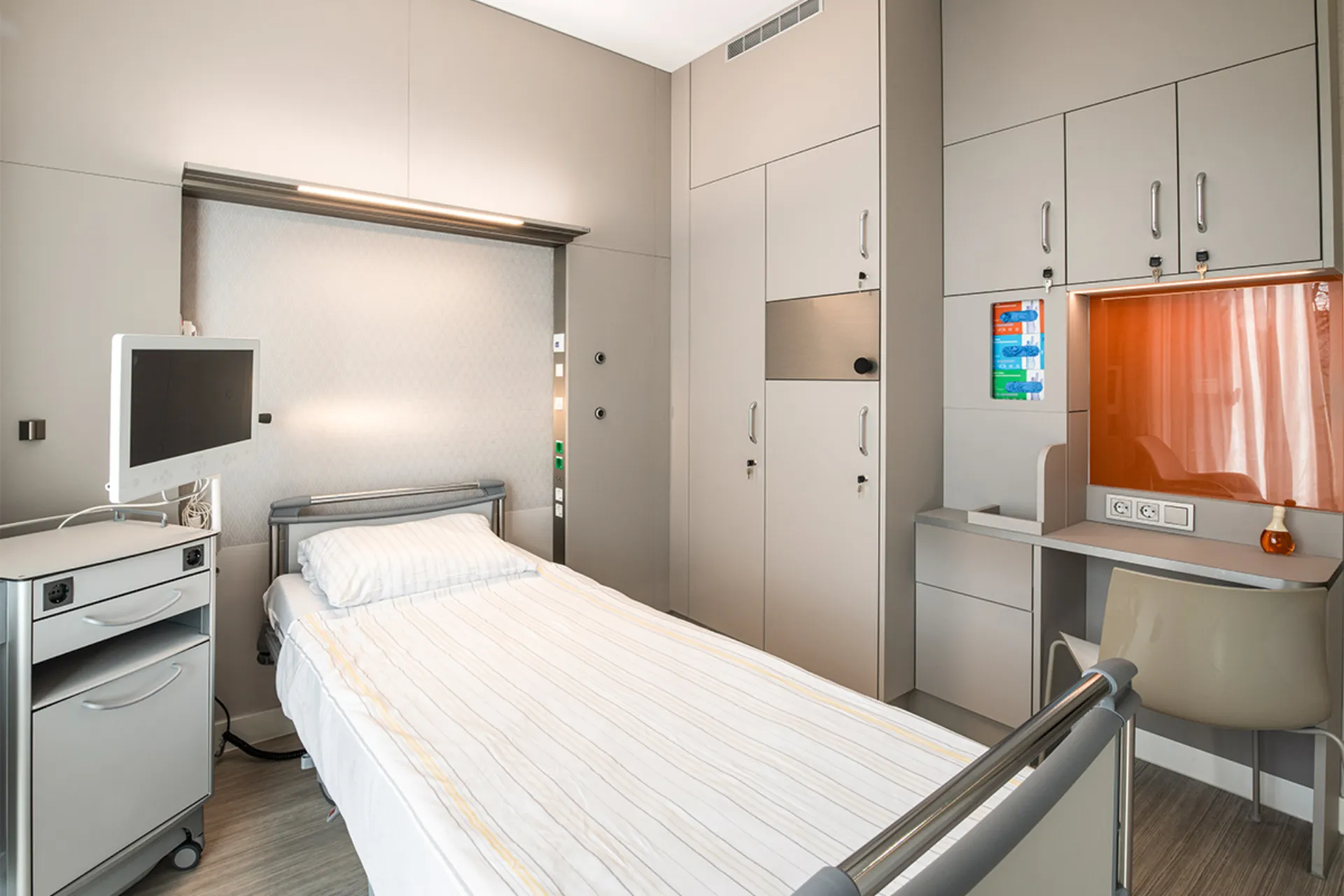 Krankenhauszimmer mit Einzelbett, Monitor auf Rollwagen, Schrankwand und Arbeitsplatz mit Stuhl und orangefarbenem Sichtschutz