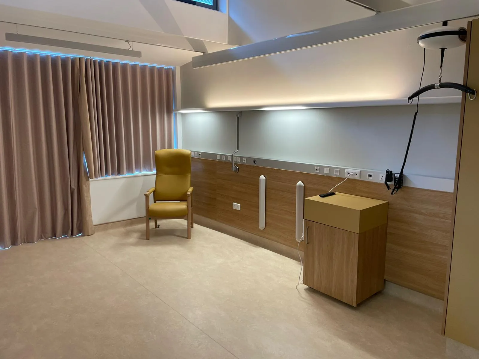 Patient room with a yellow leather armchair, wood panelling with medical gas and power connections, indirect ceiling lighting and a patient lift