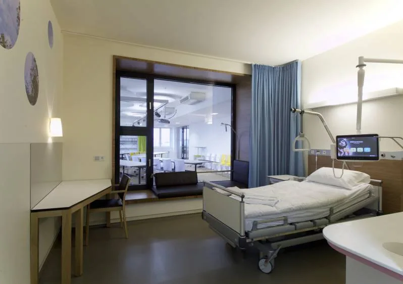 Patient room with care bed, a swivelling monitor at the bed, a wall with a circular motif and a window with a view into a bright lounge area